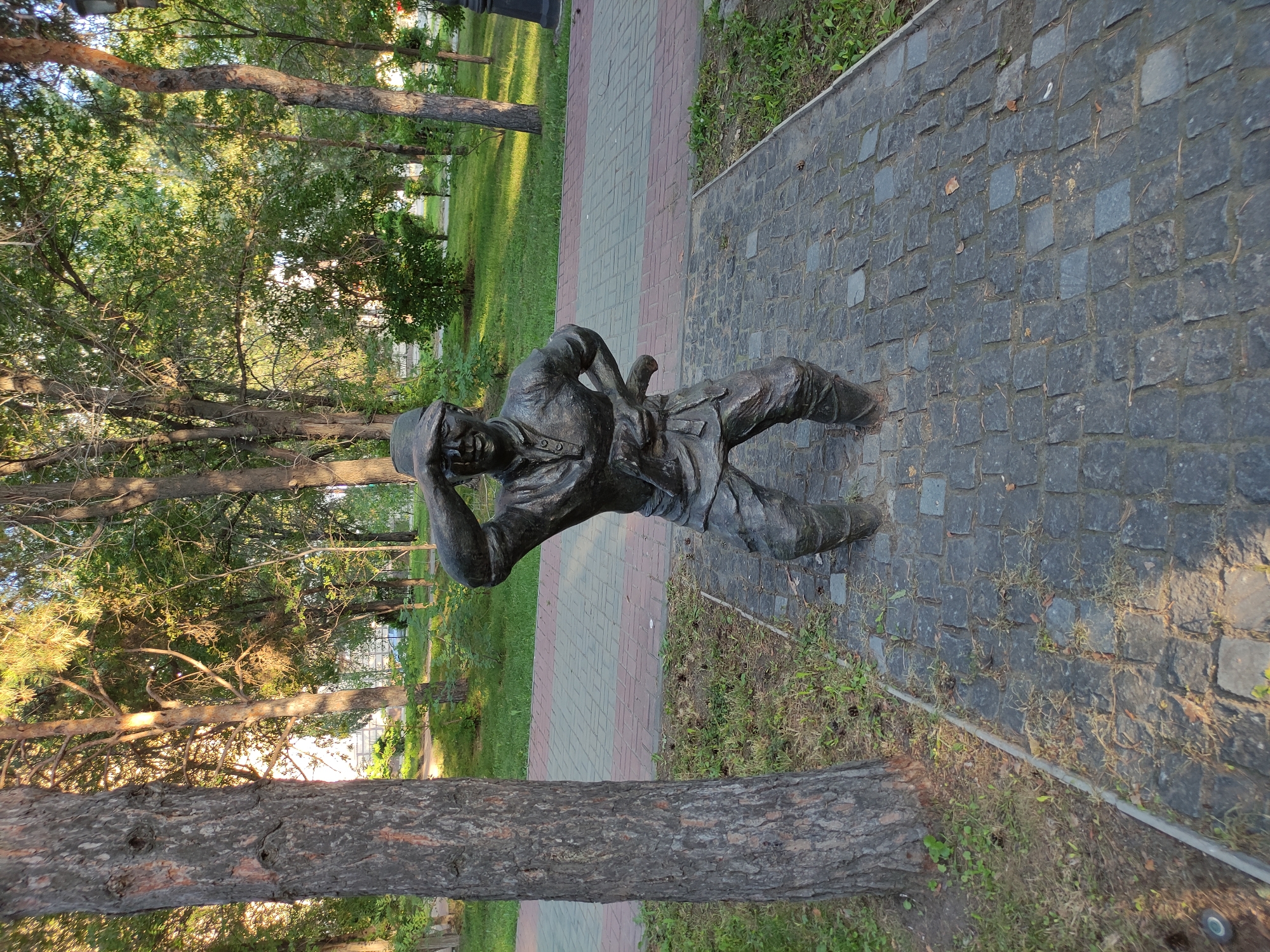 A dark bronze statue of a man in a cap and work clothes stands in a park on a cobblestone path. The man is depicted in a crouching position with his right hand raised to his forehead as if shielding his eyes from the sun. He holds a tool or weapon in his left hand. The statue is positioned in front of a paved walkway with red and grey bricks. The park has several trees and green grass. In the background, a light-colored building is visible through the trees, suggesting an urban or suburban environment. The scene is lit by natural sunlight, casting shadows on the ground. The statue's location is identified as Semey, Kazakhstan.
