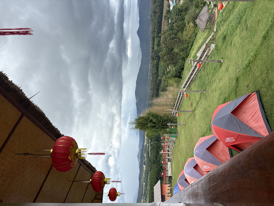 The scene depicts an outdoor elevated perspective overlooking a valley in Ban Mai Saha Sampan, Thailand, under an overcast sky. In the immediate foreground, the underside of a light-colored thatched roof structure is visible, from which three red Chinese lanterns with gold accents and red tassels are suspended. A fourth red lantern is partially visible to the left. A dark wooden railing angles across the bottom left of the frame.
The midground features a descending grassy hillside. Four red and grey/blue dome-shaped camping tents are pitched sequentially on the grass, appearing unoccupied. To the right of the tents, a series of vertically oriented wooden posts with crossbars lines the slope, each supporting a smaller red Chinese lantern. A wooden fence structure runs parallel to these posts further down the hill. A circular, open-sided gazebo with a dark thatched roof stands further down the slope to the far right.
In the background, a vast landscape extends, characterized by rolling hills covered in dense green vegetation and forested areas. Scattered buildings with orange or red roofs are discernible amidst the greenery. Distant mountains form the horizon line under a heavily clouded, grey sky, indicating potential rain. No individuals are visible.