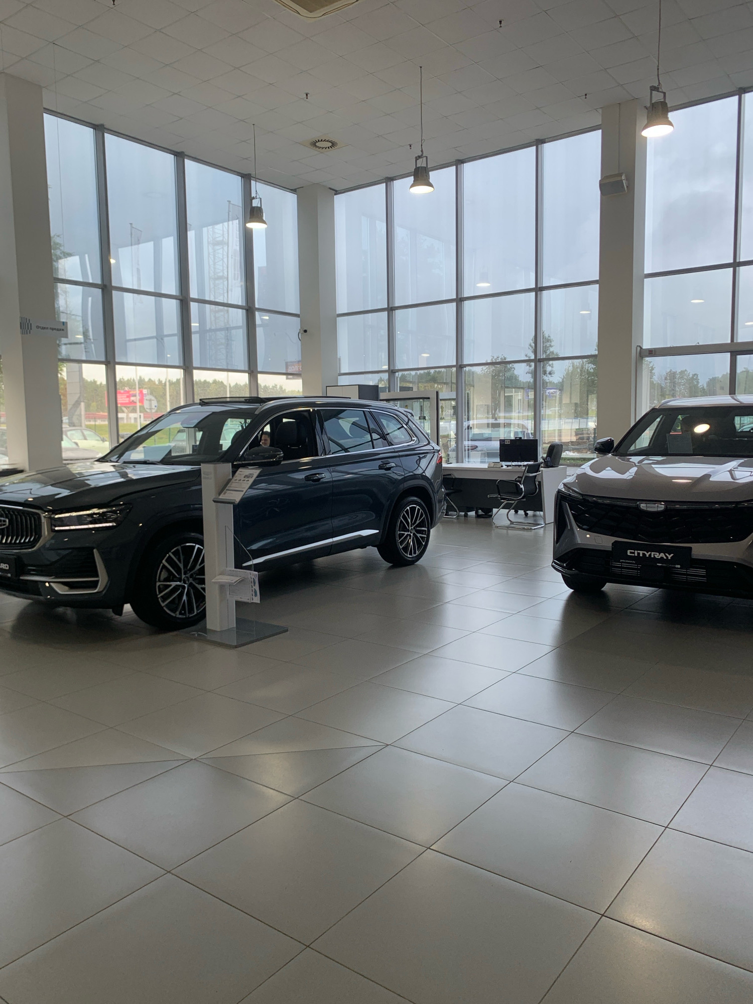 The image depicts the interior of a car dealership in Minsk, Belarus. Two cars are prominently displayed on a tiled floor. The car on the left is a dark-colored SUV, while the car on the right is a lighter-colored SUV with "CITYRAY" visible on its license plate. The dealership features a large glass facade, allowing natural light to enter the space. In the background, there are office desks and chairs, suggesting a customer service or sales area. The overall environment appears clean and modern, typical of a contemporary car showroom. The lighting in the scene is a mix of natural light from the windows and artificial light from overhead fixtures. No people are clearly visible in the immediate foreground or interacting with the vehicles.