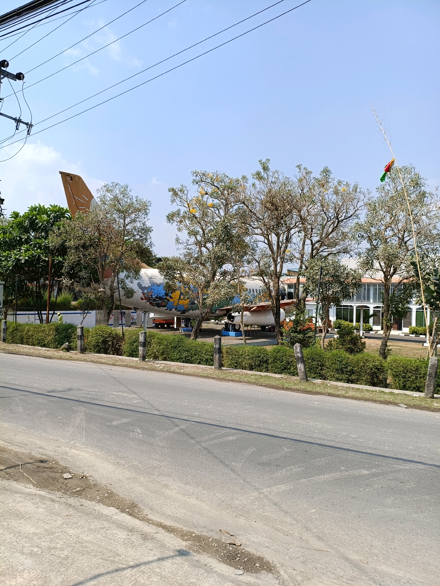 An outdoor, medium-shot, eye-level, full-color photograph shows a street in Purwomartani, Indonesia. In the foreground, a paved road is visible, with tire marks and a dirt and debris-covered shoulder on the left. Behind the road, a low, trimmed hedge runs horizontally across the frame. Beyond the hedge, a retired airplane is on display, positioned on a grassy area. The plane is white with brown and blue accents and features cartoon characters painted on its side. Several trees, some with sparse leaves and yellow blossoms, are interspersed between the viewer and the airplane. In the background, a modern building with large windows is visible behind the airplane. A tall, thin pole with a small, colorful flag is planted in the ground on the right side of the frame. Overhead, power lines crisscross a bright blue sky with a few wispy clouds. The overall scene suggests a public space or museum showcasing the aircraft.