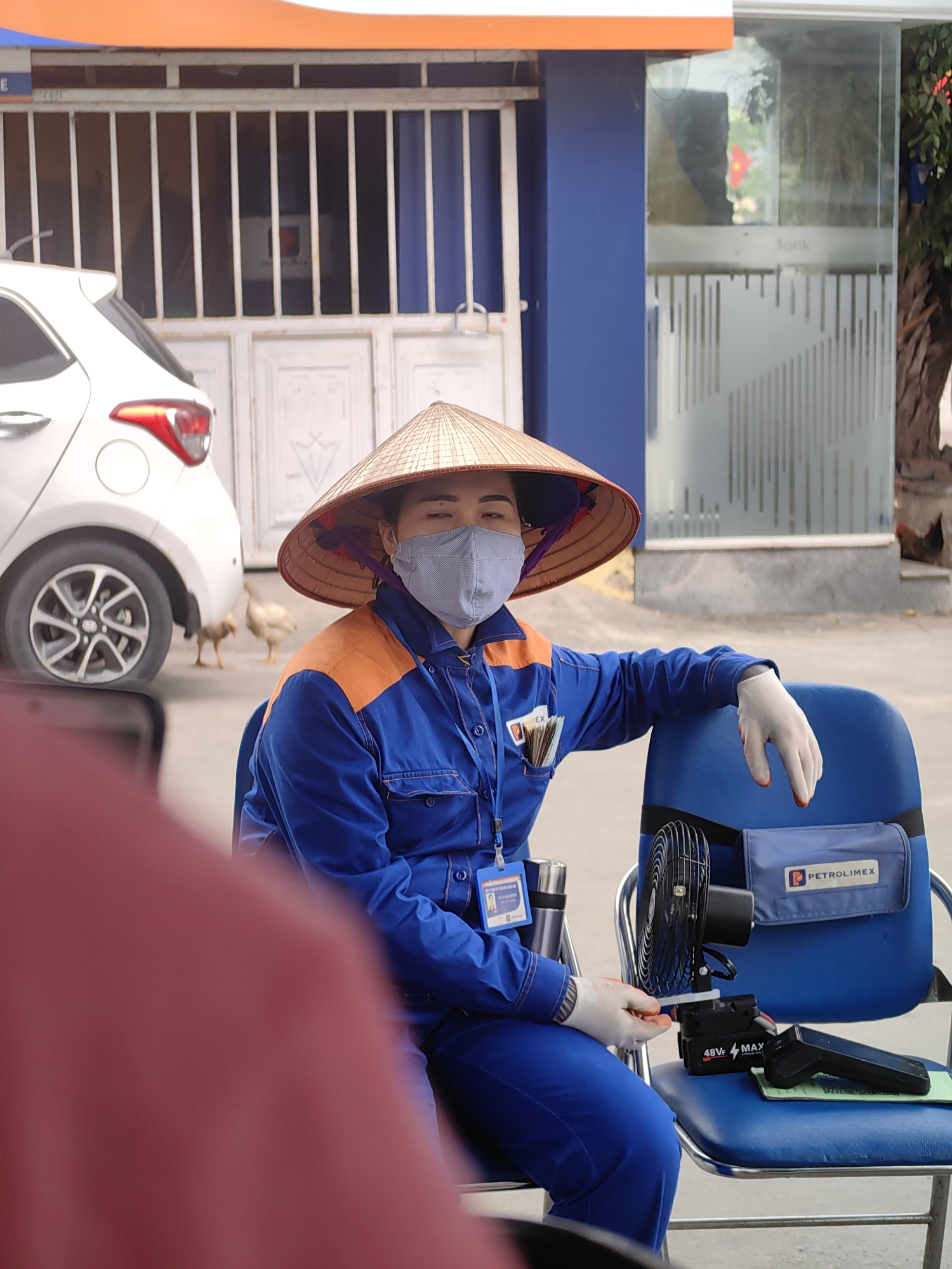 A woman wearing a traditional Vietnamese conical hat (nón lá), a light grey face mask, and a blue and orange uniform is seated on a blue chair. She is also wearing white gloves and has an ID badge visible on her uniform. Her left arm rests on the back of another blue chair beside her. Money is tucked into her uniform's chest pocket.
On the adjacent chair, several items are placed: a small portable black fan, a battery pack or power bank labeled "48Vf MAX", a dark handheld device (possibly a payment terminal or scanner), and a blue pouch with the "PETROLIMEX" logo. A thermos or water bottle is also visible near her, attached to her belt or another chair leg. Part of a red smartphone is in the lower left foreground, slightly obscuring the view.
The scene appears to be outdoors during daytime, with bright natural light. In the background, a white car is parked. Two light-colored chickens are foraging on the ground behind the car. A building with white grated windows and a blue wall is visible, along with a glass entrance door that has faint patterned frosting and a partially visible word "Bank". An orange architectural feature or awning is at the top of the frame. The woman's calm posture, the uniform, and the "Petrolimex" branding suggest she might be a staff member at a service station or similar establishment, possibly on a break. This setting is located in Cao Xanh Ward, Vietnam.