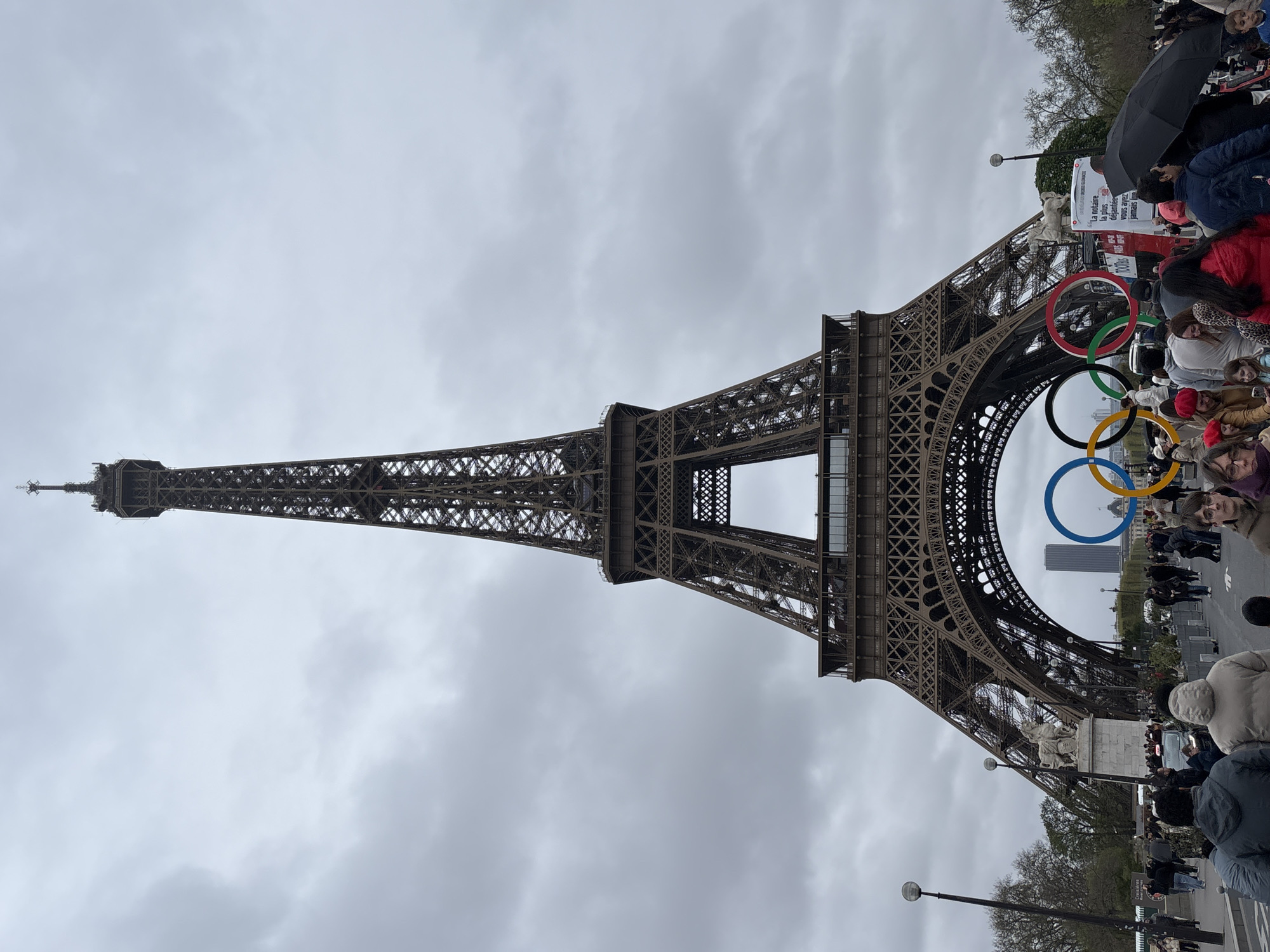 The image shows the Eiffel Tower in Paris, France. The tower is prominently displayed with the Olympic rings affixed to the tower's base. There is a crowd of people gathered around the base of the tower. The sky is overcast.