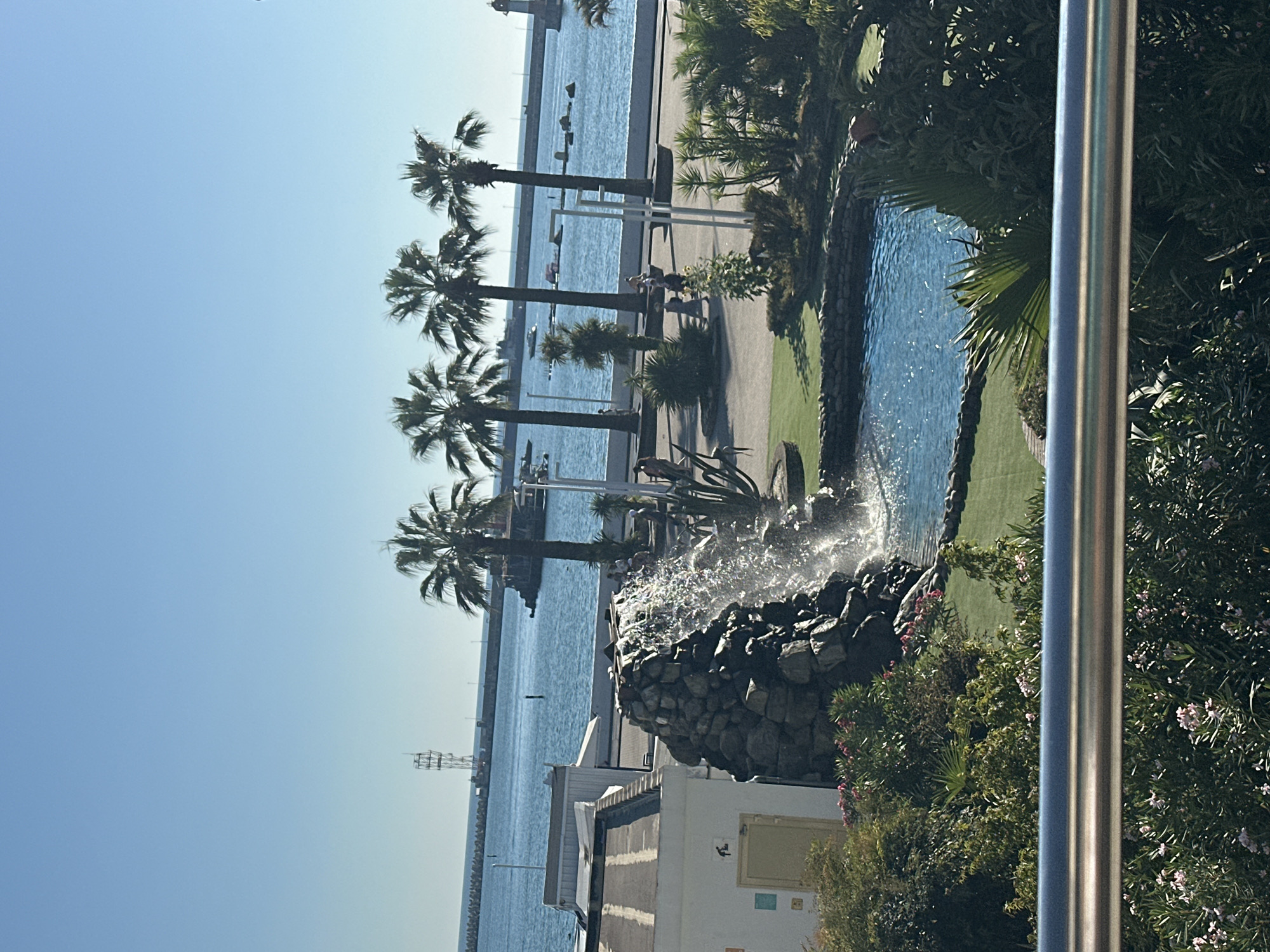 A clear, sunny day unfolds in Sochi, Russia, with a view of the Black Sea and a vibrant man-made waterfall. In the foreground, lush green foliage and a metal railing frame the scene. The waterfall, constructed from dark rocks, cascades into a bright blue pool, surrounded by meticulously manicured green grass. Beyond the waterfall, a paved walkway lines the waterfront, dotted with several palm trees that stand tall against the azure sky. People stroll along the walkway, some walking alone, others in small groups. A white building with a light-colored door is visible to the left of the waterfall. In the distance, the Black Sea stretches out, with a breakwater, a lighthouse, and a tall radio tower marking the horizon. Boats can be seen in the water, suggesting a bustling port or harbor.