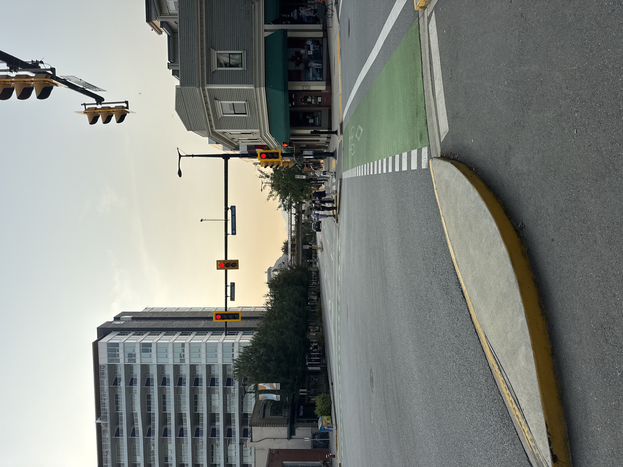 The image depicts an intersection in North Vancouver, Canada, likely near Lonsdale Quay, as indicated by a street sign. A tall, modern apartment building with many balconies stands on the left, while a historic-looking building with a green awning occupies the right side of the intersection. A bright green bike lane with a white bicycle symbol is visible, crossing the intersection. Traffic lights are red, indicating a stop. Several pedestrians are present on the sidewalk, some standing and others walking. The sky is overcast with a hint of a sunset glow. The overall scene is a typical urban streetscape with a mix of contemporary and older architecture.