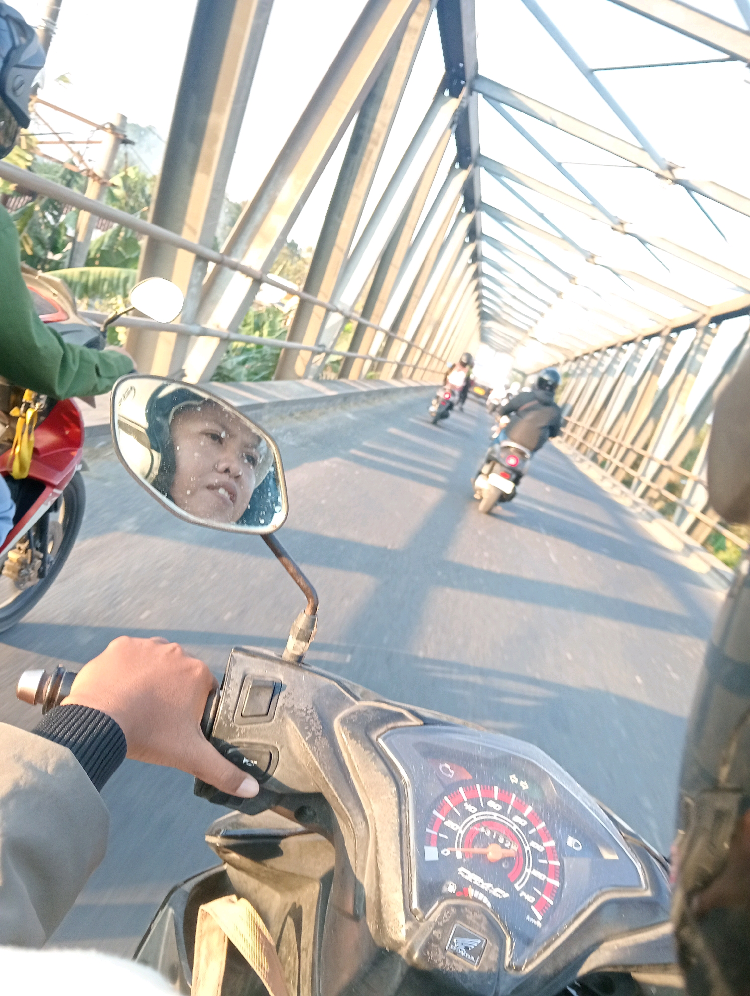 The image captures a POV shot from a motorcycle traveling across a bridge. The motorcycle has a black and red dashboard with a speedometer and various indicator lights. The rider's hand, wearing a dark gray jacket with ribbed cuffs, is visible on the handlebars. A side mirror reflects the rider's face, which is partially obscured by a helmet. In the background, several other motorcycles are also on the bridge, heading in the same direction. The bridge is an elevated structure made of steel beams, and the surrounding environment appears to be a tropical landscape with lush green vegetation. The lighting suggests it is either early morning or late afternoon, with the sun casting long shadows. This bridge is identifiable as Jembatan Sosrodilogo in Bojonegoro, Indonesia, a prominent landmark known for its length and the scenic views it offers.