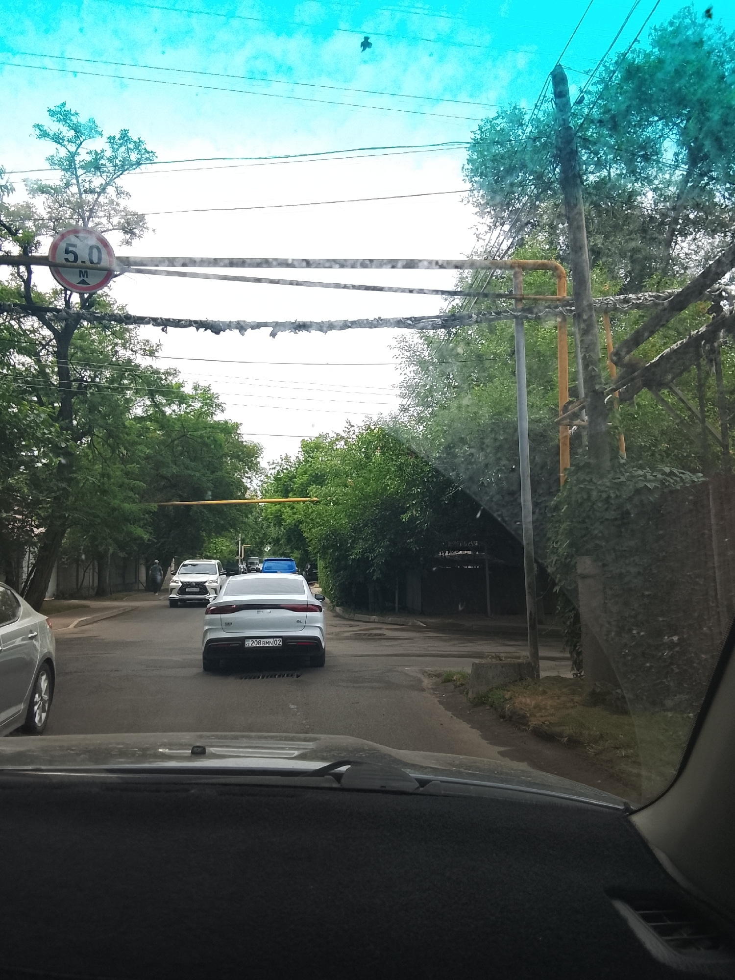 The image depicts a street scene from inside a vehicle, likely in Almaty, Kazakhstan. The perspective is from the driver's seat, looking forward through the windshield, which has some smudges and streaks. The time of day appears to be daytime, with bright, overcast skies. The weather seems pleasant, possibly warm, as indicated by the lush green trees lining the road.
The street is paved and appears to be a residential or suburban area. Several cars are visible. Directly ahead, a white sedan with a license plate reading "208 BMN 02" is the most prominent vehicle. Further ahead, another white SUV is turning, and behind it, a blue vehicle is partially visible. To the left of the road, there's a fence and more trees. On the right, utility poles and more foliage create a dense green barrier.
A prominent circular road sign with a red border and a white interior is visible on the left, displaying "5.0" and "M" with an arrow pointing downwards, indicating a height restriction of 5.0 meters. The overhead wires are strung between poles and appear somewhat disheveled. In the distance, there's a structure that looks like a covered walkway or parking area. The overall impression is a quiet, everyday scene on a slightly cloudy day.