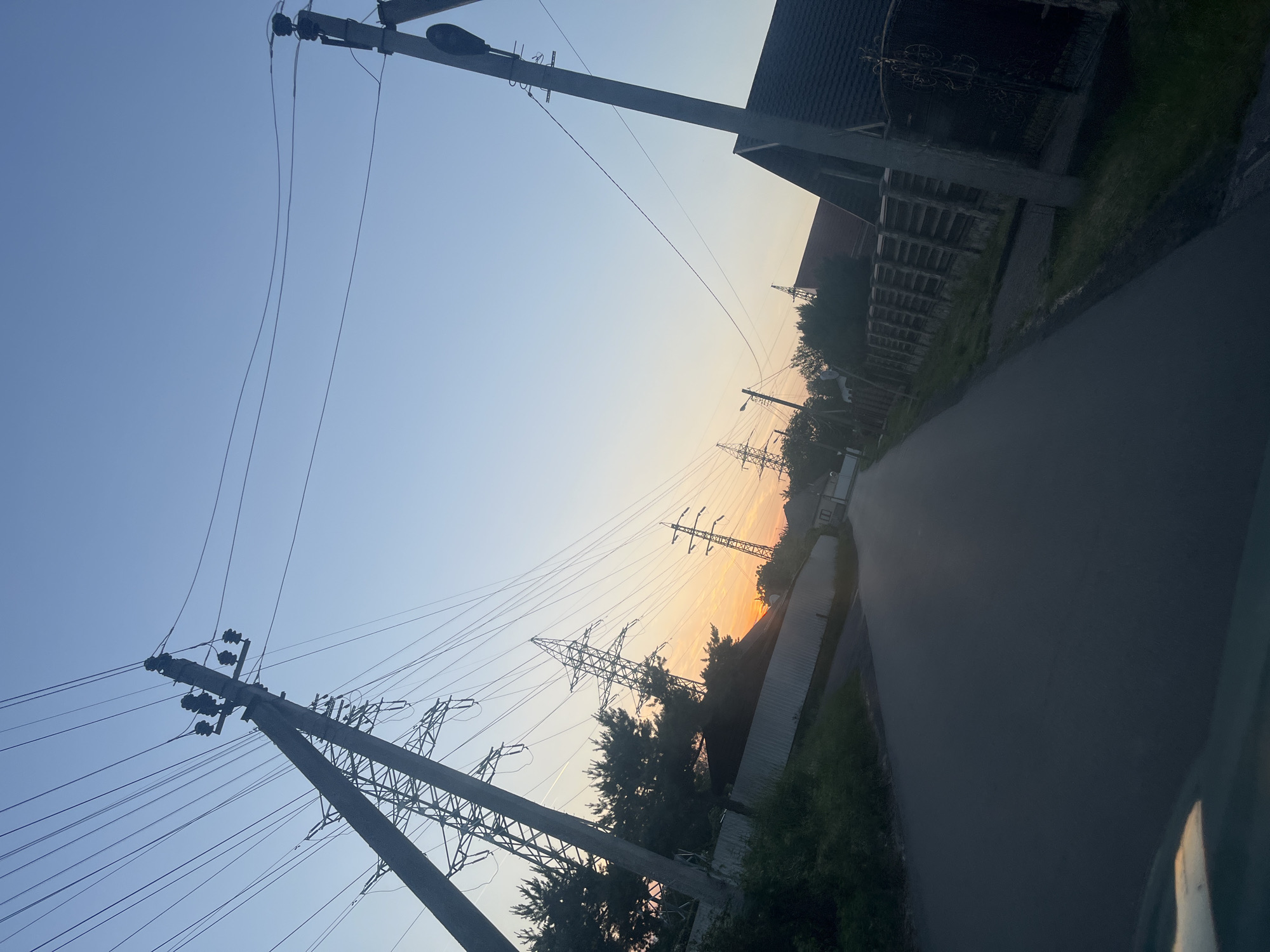 The image depicts a rural or semi-urban landscape at either sunrise or sunset in Storozhnytsia, Ukraine. The sky, clear and cloudless, dominates the upper portion of the frame, transitioning from a light blue overhead to a warm orange and yellow glow on the horizon, indicating the low angle of the sun.

Prominent in the scene are numerous electricity infrastructure elements. Several dark utility poles, some with attached streetlights, stand in the foreground and midground, carrying a dense network of power lines that crisscross the sky. Further in the distance, large, angular metal transmission pylons are silhouetted against the colorful sky, suggesting a significant power grid in the area.

A paved road stretches from the lower right of the image, curving gently into the distance. On both sides of the road, dark, silhouetted buildings with sloped roofs and fences are visible, nestled amidst trees and bushes that also appear dark against the bright sky. No people or vehicles are visible, contributing to a quiet and tranquil atmosphere. There is no visible text in the image.