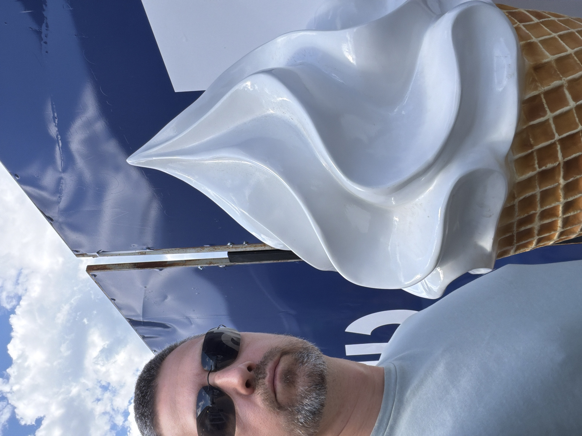 A man wearing sunglasses and a light blue shirt stands in front of a large, three-dimensional ice cream cone advertisement. The ice cream cone is predominantly white with a brown waffle cone. The advertisement features a blue background with white text or shapes. The image was taken outside during daylight under a sky partially covered by white clouds.