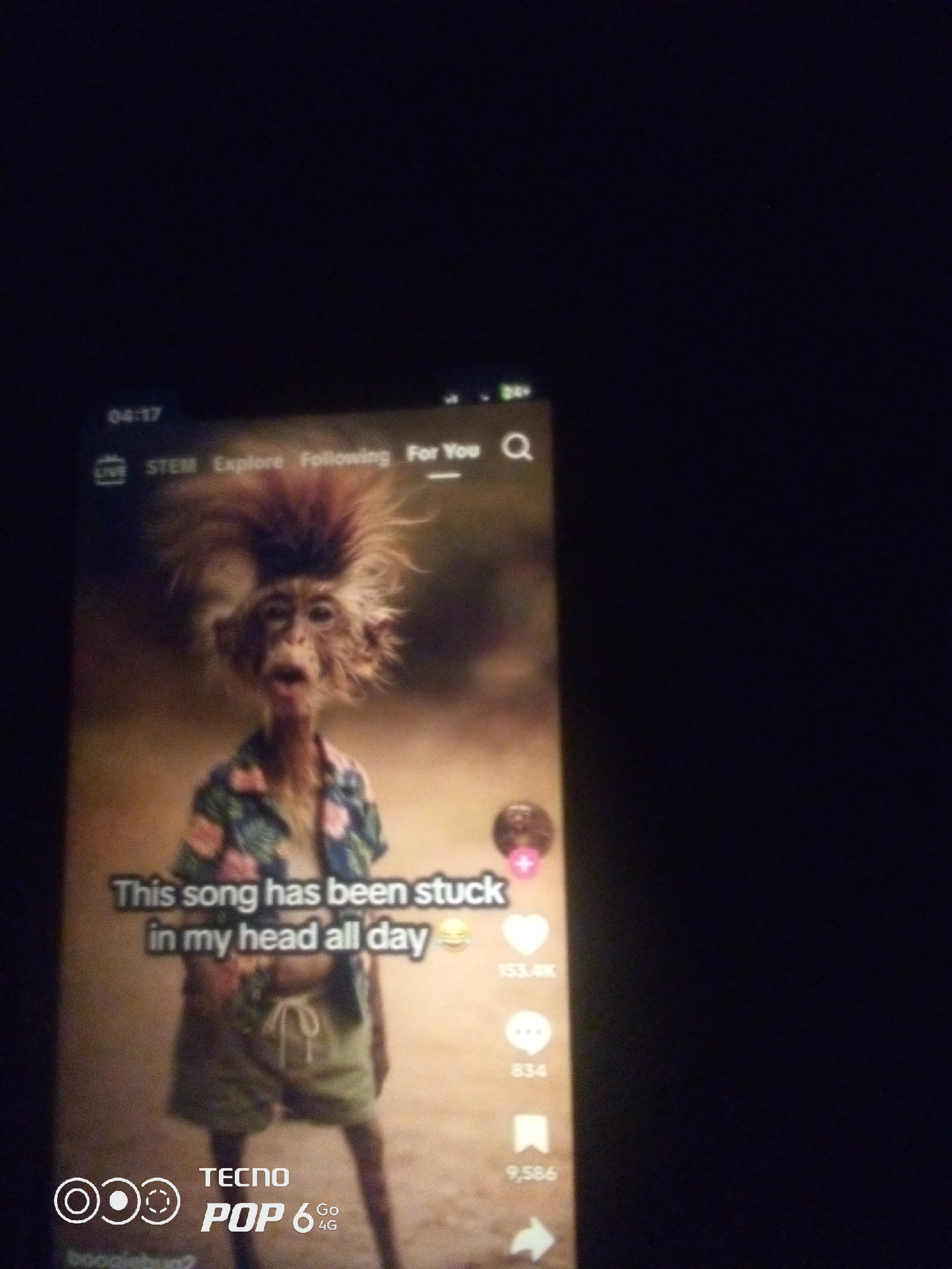 The image shows a TikTok video on a phone screen. The video features a monkey with spiky hair wearing a Hawaiian shirt and shorts. Text on the screen reads "This song has been stuck in my head all day". Social media interaction icons and counts are visible on the right side of the screen. The bottom of the phone screen shows the TECNO POP 6 4G logo. The phone is on a dark background.