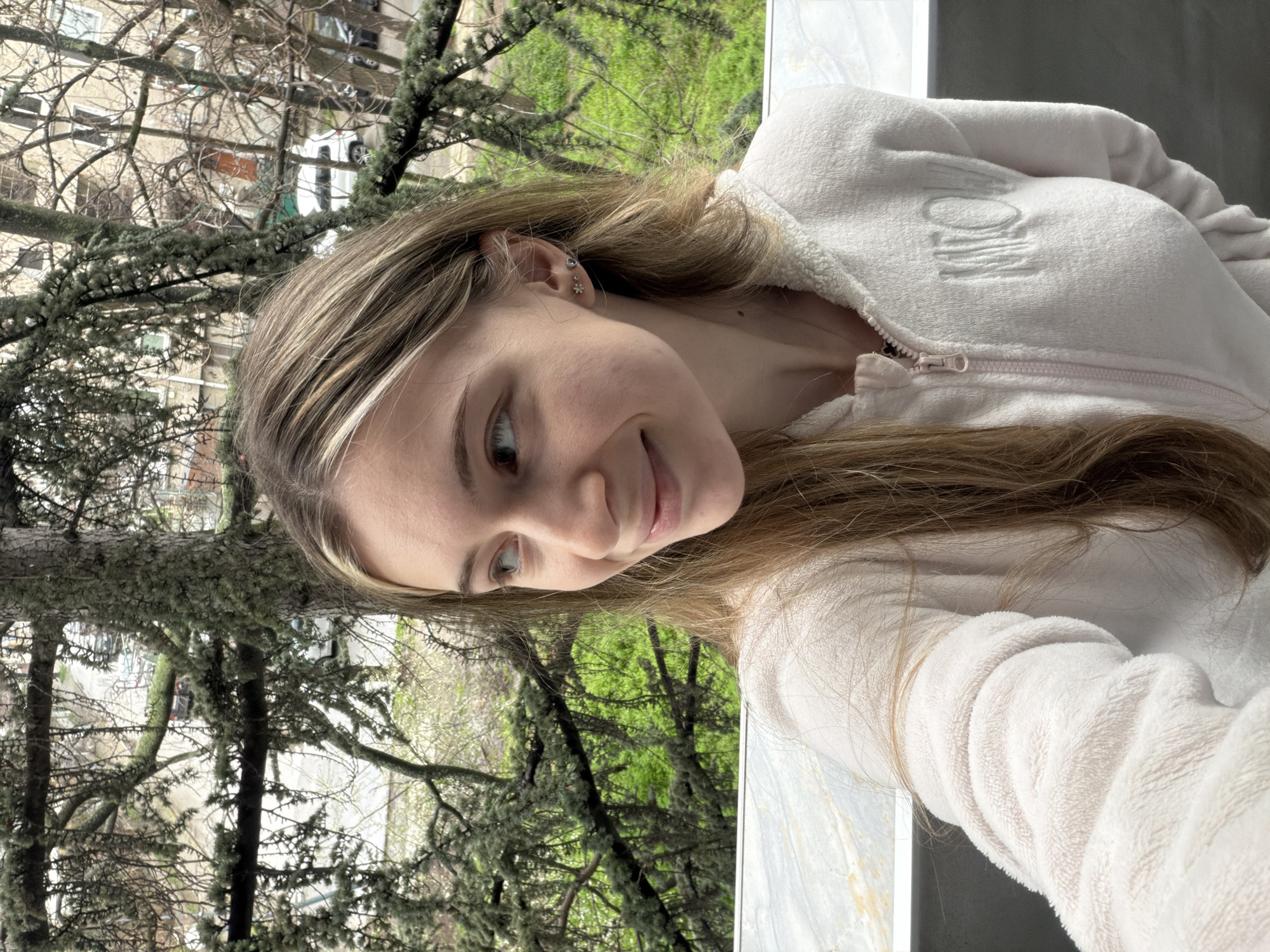 A young woman with light brown hair and a single blonde streak near her face is pictured in a selfie. She's wearing a soft, light pink zip-up jacket. Her left ear is visible, adorned with multiple earrings. The background shows an urban landscape through a window, with leafless trees and buildings. Some greenery is visible in the distance. The location may be near the Plovdiv cityscape, however there is not enough information to identify a specific site in the city.