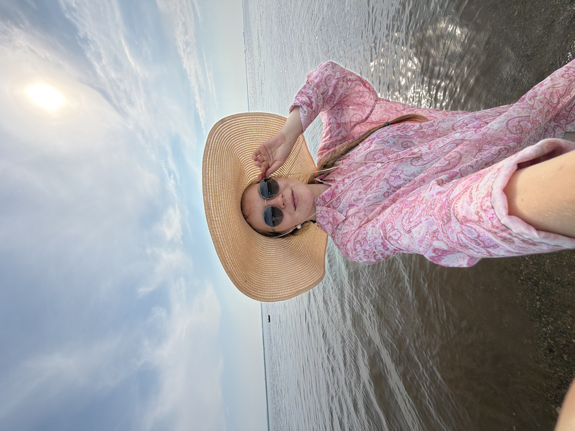 A young woman wearing a large straw hat and a pink patterned shirt stands in shallow water, likely a lake or river, with a bright sun in the sky overhead. She is holding the brim of her hat with one hand and is looking towards the camera. Her hair is styled in a braid. The water is calm with ripples reflecting the sunlight. In the distance, there is a very small boat on the water. The location is the city of Krasny Bor, Russia.