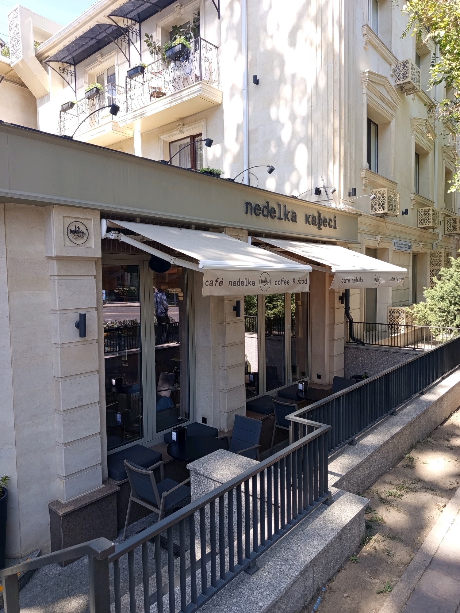 The image depicts the exterior of a cafe named "Nedelka Cafe" located on Zhibek Zholy Street in Almaty, Kazakhstan. The cafe features a light-colored stone facade with prominent architectural details, including decorative cornices and columns. A beige awning extends over the entrance and windows of the establishment, providing shade. The cafe's name, "nedelka кафесі" (nedelka cafe in Kazakh and Russian), is displayed in dark lettering above the awning. Several windows with dark frames are visible, revealing glimpses of the interior where tables and chairs are arranged.
In front of the cafe, a small outdoor seating area is set up with dark tables and chairs, separated from the sidewalk by a metal railing. Beyond the railing, a sloping path leads down to a ground-level area with patchy grass and dirt, indicating a sidewalk or pedestrian walkway. The building itself appears to be multi-story, with balconies adorned with planters on the upper floors. The overall impression is of a modern, well-maintained urban commercial establishment. The time of day appears to be daytime, with shadows cast by the sun suggesting it is mid-morning or afternoon.