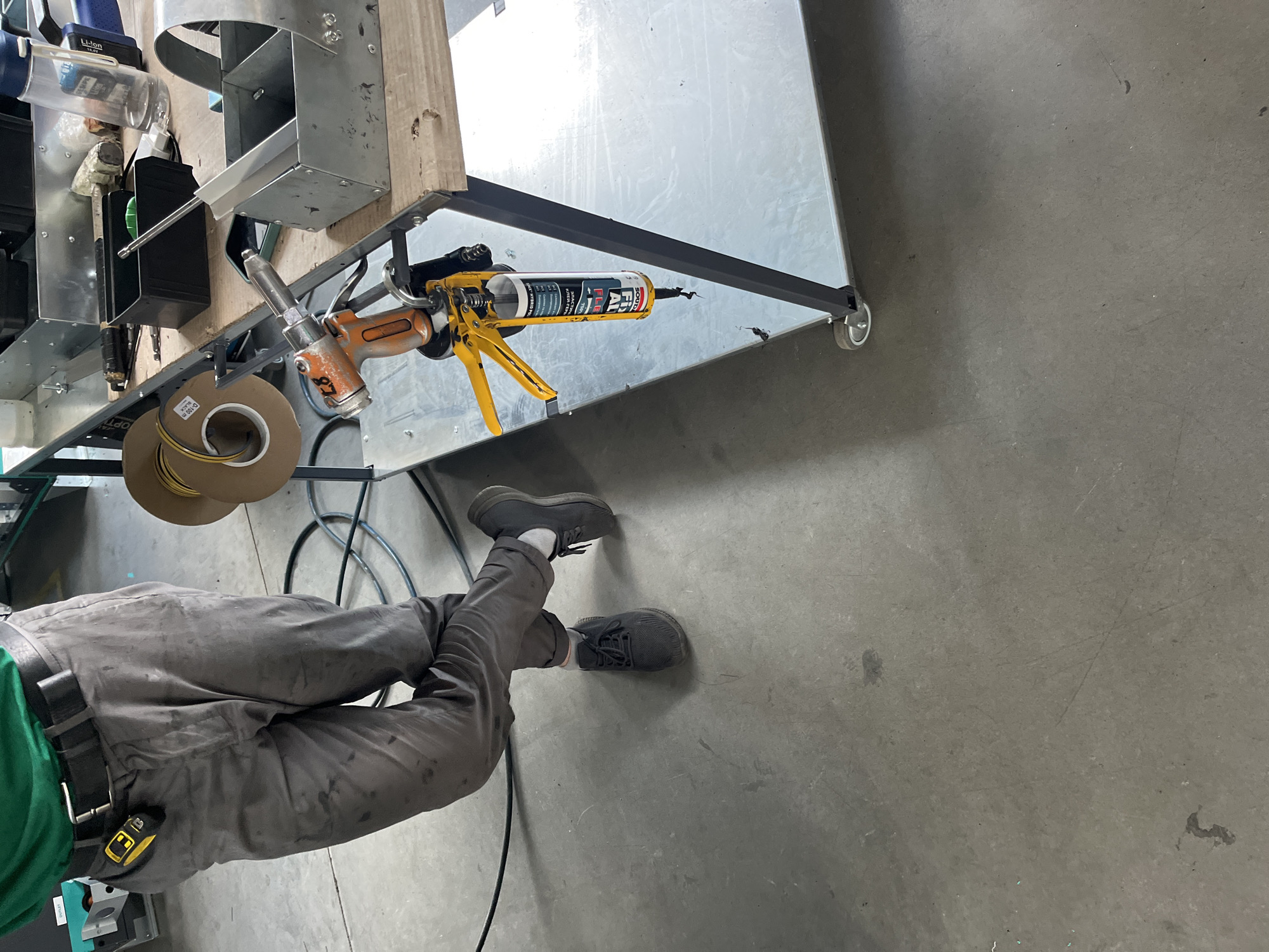 The image shows a person wearing a green shirt and grey work pants, standing in what appears to be a workshop or industrial setting. The person is wearing a black belt with a yellow tape measure clipped to it. Their left leg is crossed over their right, and they are wearing black sneakers. In the background, there are various tools and materials, including a caulking gun with a tube of sealant, a spool of tape, and metal containers. The floor is concrete. The scene is not located in Kyiv, Ukraine.
