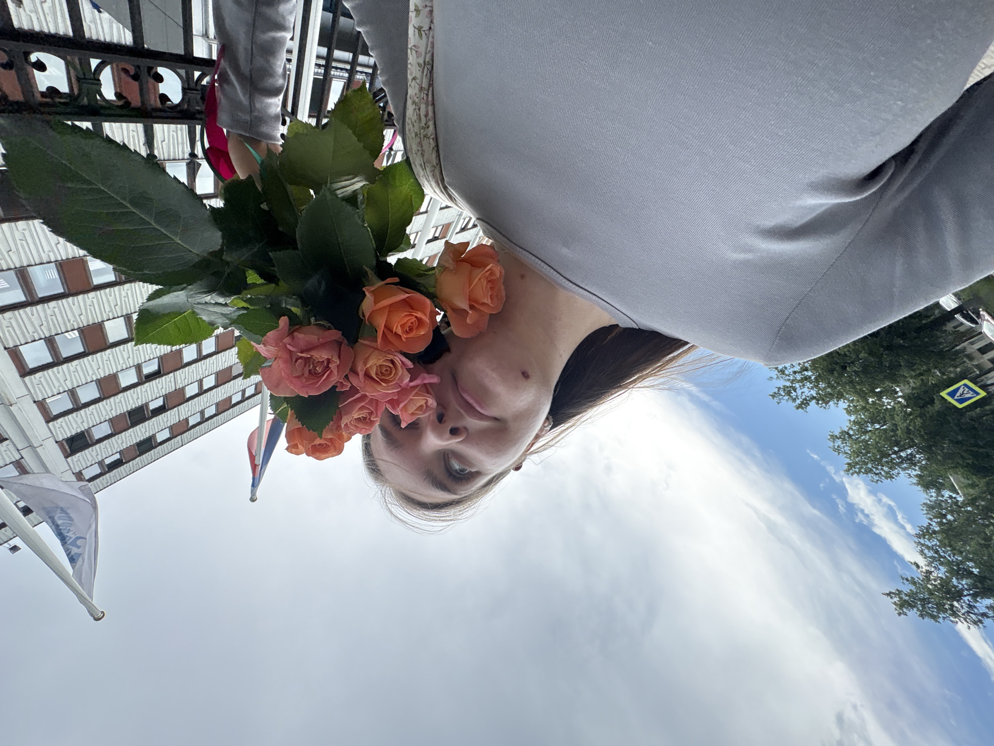 A young woman with light brown hair is holding a bouquet of pink and orange roses to her face. She is wearing a gray long-sleeved shirt. In the background, a building with white and brown brickwork and rows of windows is visible, along with a cloudy sky and trees. The image appears to have been taken outdoors on a bright day, possibly in the city of Chaikovsky, Russia, as indicated by the presence of flags and a street sign.