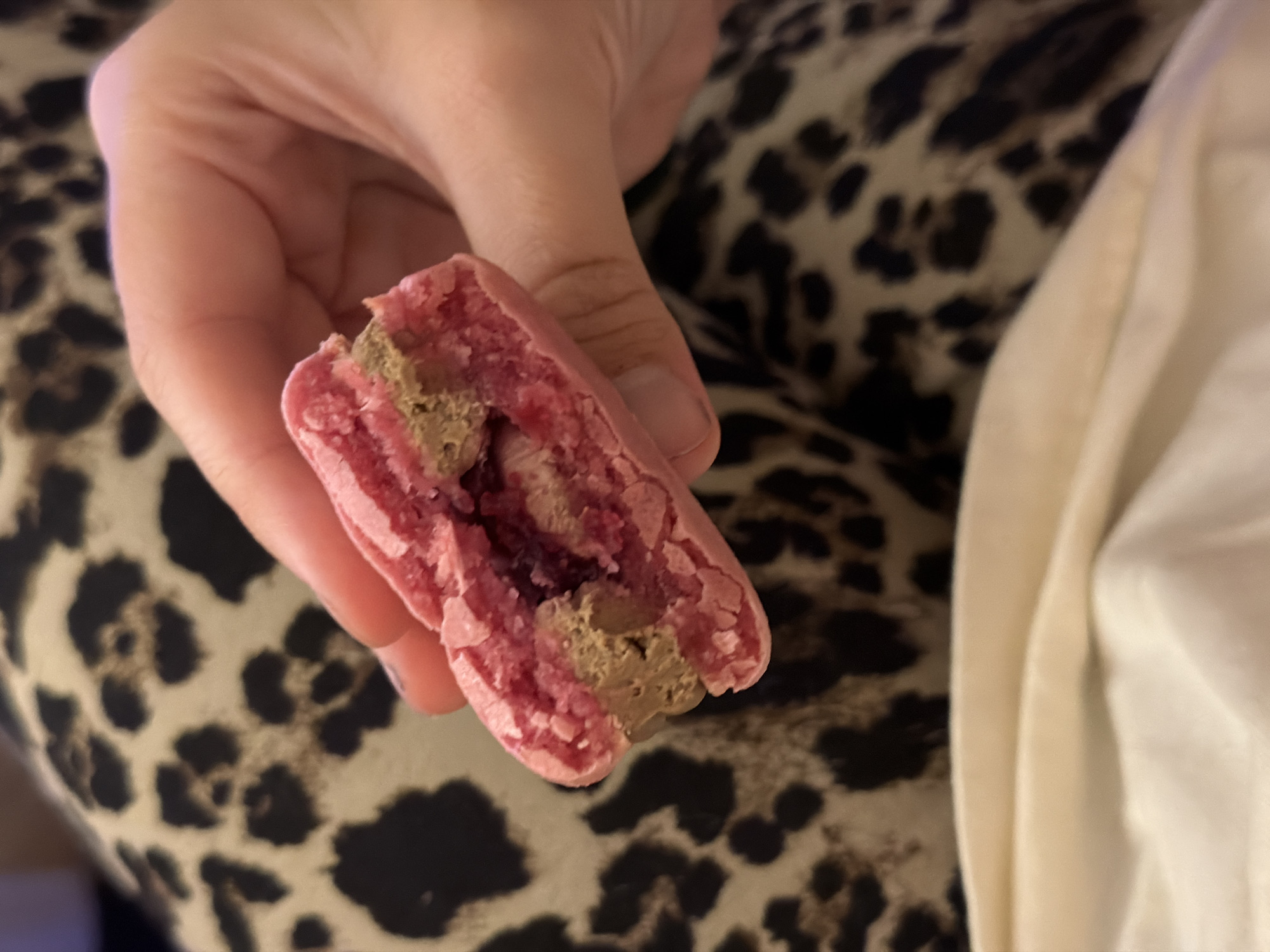 A partially consumed pink macaron is held in a human hand. The filling appears to be a combination of light and dark brown textures. The background features a surface patterned with brown and black spots, possibly animal print fabric. The specific location in Tel-Aviv, Israel cannot be determined from this image.