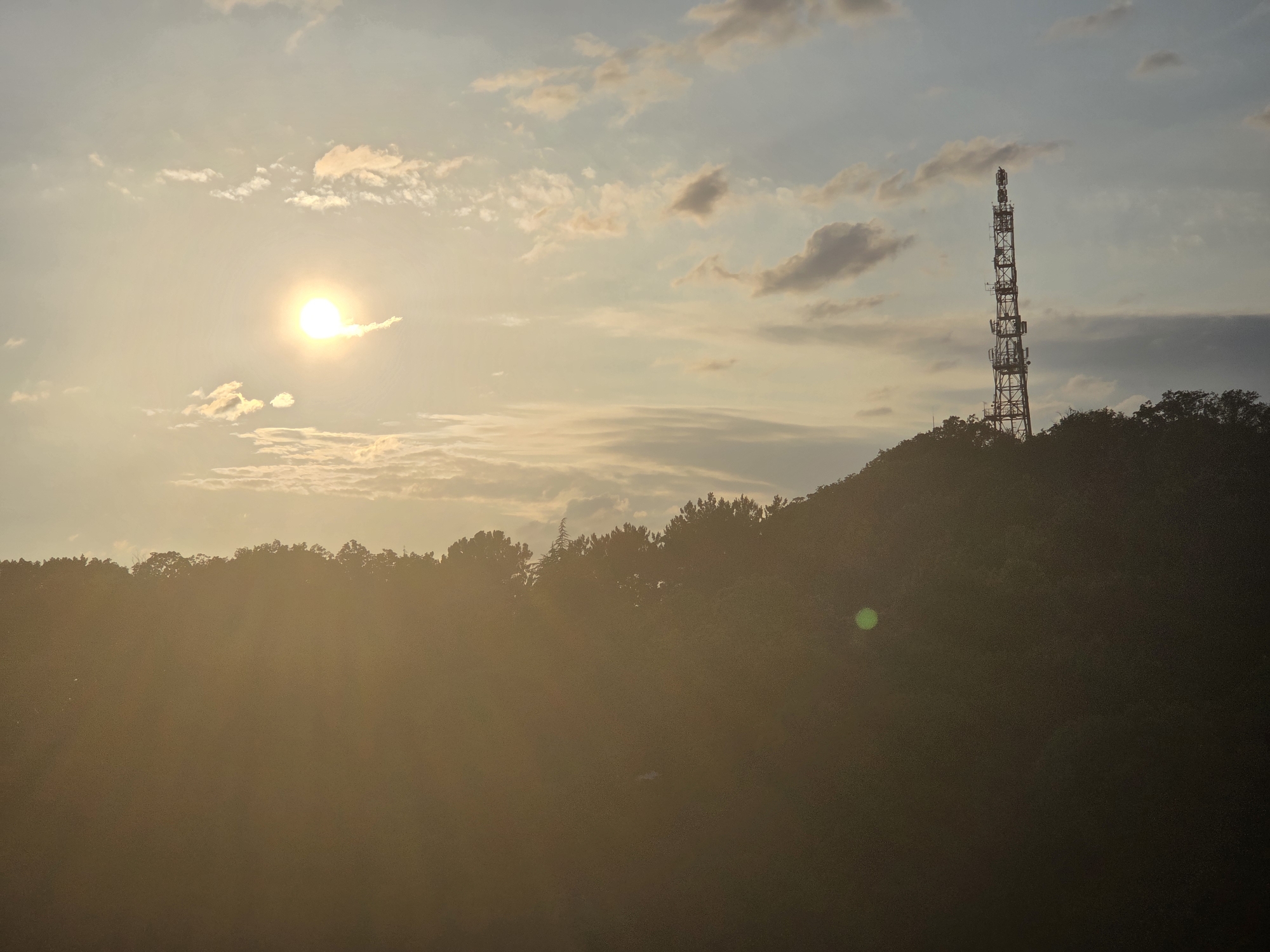 An outdoor, wide shot captures a silhouette of a cell tower on a hill against a cloudy sky at sunset. The sun is a bright orb in the upper left quadrant, casting a warm glow and lens flare across the scene. The cell tower is tall and latticed, positioned on the right side of the frame, atop a dark, tree-covered hill. The foreground is dominated by the hazy, sun-drenched foliage of the hill, obscuring any fine details of the landscape below. The sky is a gradient of pale blues and yellows, with scattered clouds tinged orange by the setting sun.