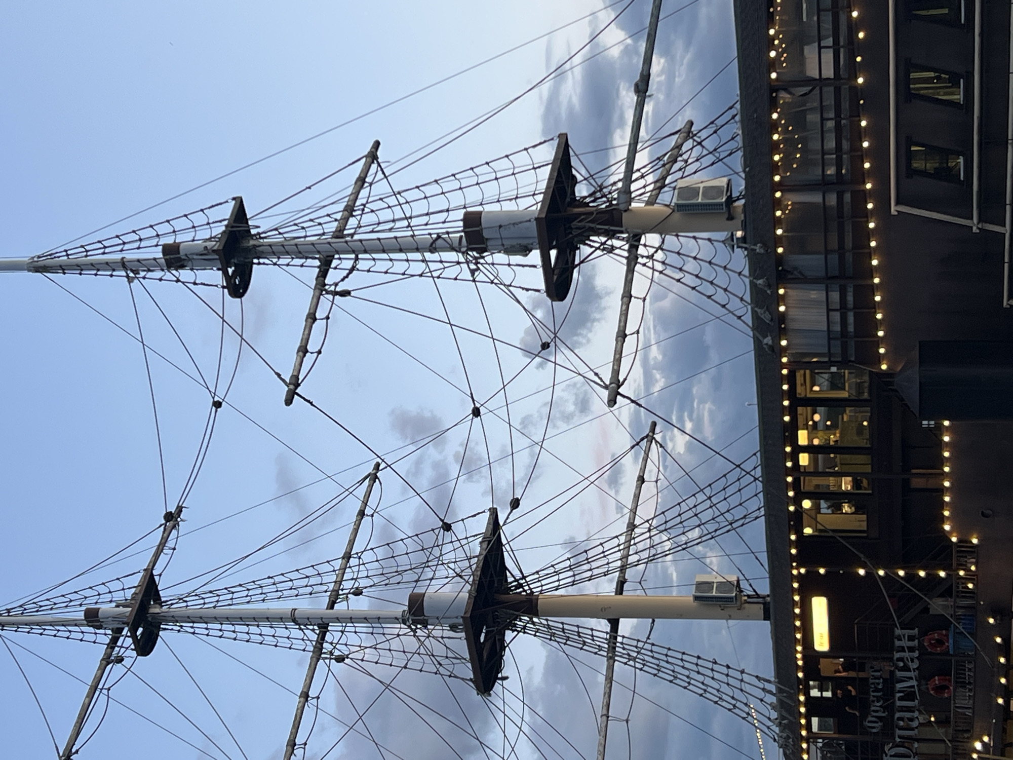 The image shows a detailed view of the masts and rigging of a tall ship, likely docked or as a permanent structure. The ship's hull is visible in the lower portion of the image, featuring windows and decorative string lights. Text is visible on the hull, reading "Фрегат ФЛАГМАН" (Frigate FLAGMAN). The sky above is a mix of blue with scattered clouds, suggesting it is either dawn or dusk. The scene is set in the city of Veliky Novgorod, Russia, and the "Frigate Flagman" is a restaurant located on the Volkhov River embankment.
