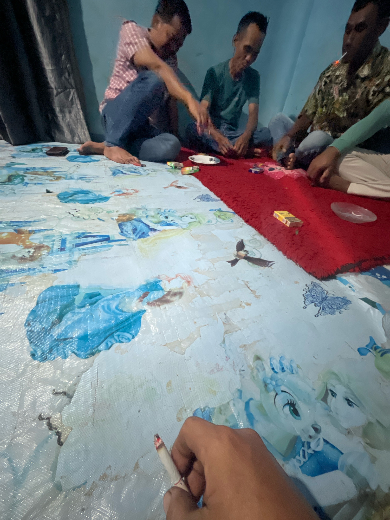 Four men are sitting on the floor in what appears to be a room in Kebumen, Indonesia. The floor is covered with a tarp featuring Disney characters, and a red rug is laid out on top of it. The men are engaged in an activity, possibly playing a game, with some items spread out on the rug, including a pack of cigarettes and a small plate. One man in the foreground is holding a lit cigarette. The lighting suggests it is nighttime or the room is dimly lit. The overall impression is a casual gathering in a domestic setting.