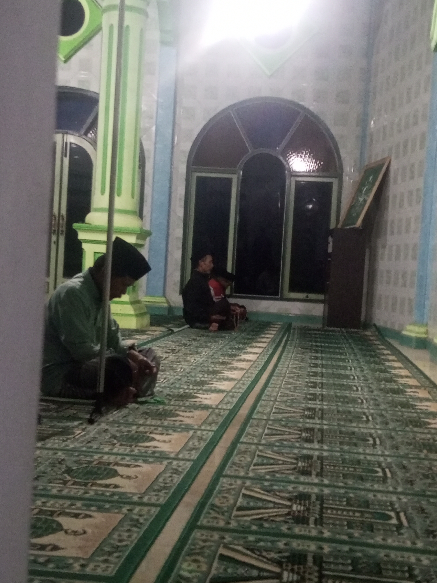 The image shows the interior of a prayer room, likely a mosque, in Andongsili, Indonesia. Two individuals are seated on a green and beige prayer rug. The person in the foreground, on the left, is wearing a light green shirt and a black peci (cap). They are seated with their legs folded and hands resting on their lap, appearing to be in prayer or contemplation. To the right and slightly behind, another individual, wearing darker clothing, is also seated. The room features patterned walls, possibly tiled, in light and blue colors. A large arched window with dark panes is visible in the background. Green and white striped columns are present, supporting the ceiling. A tall, dark brown cabinet and a framed object leaning against it are also visible. The floor is covered with a prayer rug featuring intricate green and beige geometric patterns. The lighting is dim, with some light sources contributing to bright spots in the upper part of the image.