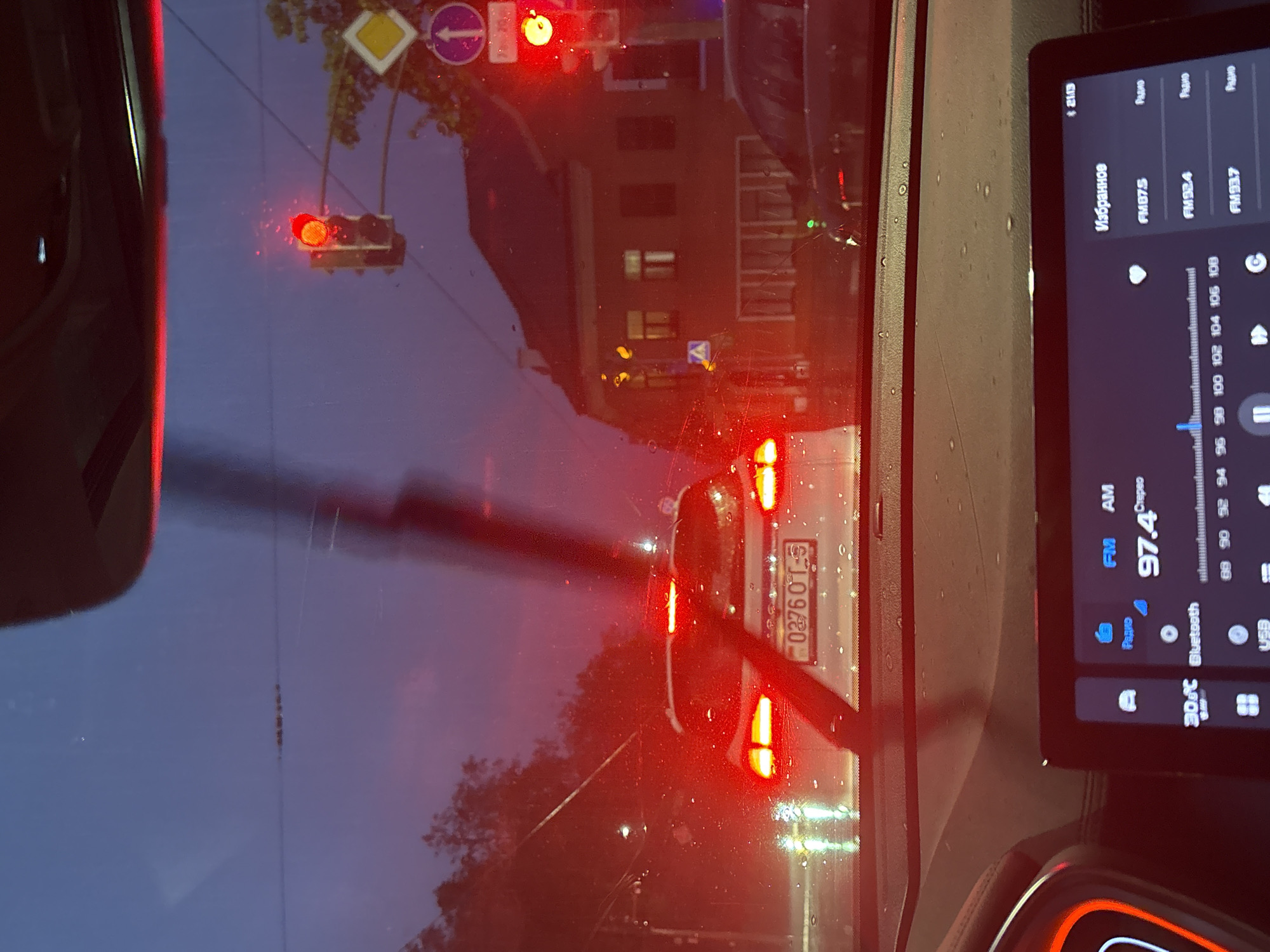The image shows a view from inside a car, looking out through the windshield at a street scene. It appears to be dusk or night, and there's light rain or moisture on the windshield.
A red traffic light is visible. Street signs, including a diamond-shaped yellow warning sign and a blue circular sign with a white arrow, are also seen. A building is visible in the background.
The focus is on a white car directly in front, its red tail lights illuminated. The license plate number "03760T-5" is partially visible on the rear of the car.
Inside the car, a digital screen displays radio information and temperature. The radio station is at 97.4 FM, and the temperature is 30°C. The screen is displaying Cyrillic text.
The license plate is a Belarusian format license plate, and Cyrillic on the display indicates a location in Belarus. Based on the radio station displayed, the location could be in Minsk, Belarus.