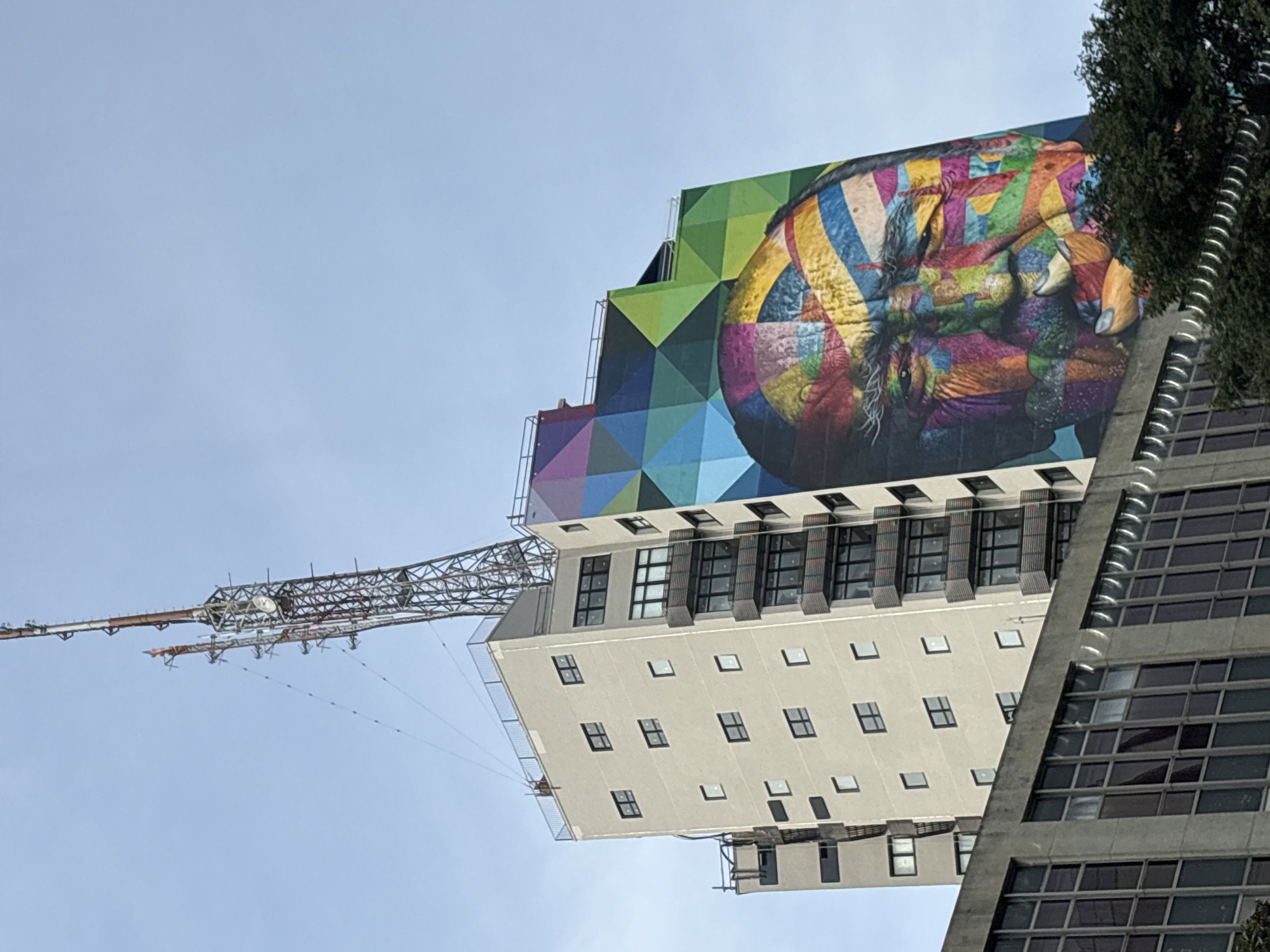The image captures a towering telecommunication antenna juxtaposed against a modern, light-colored building adorned with a vibrant, geometric mural of a man's face. The mural, painted with bold strokes of various colors including red, yellow, green, blue, and purple, covers a significant portion of the building's facade, creating a striking visual contrast with the more subdued architecture. The antenna, a complex lattice structure of metal, extends high into the clear blue sky, suggesting it is a prominent feature of the urban landscape. The building itself features numerous windows and balconies, indicating its use as a residential or office complex. The lower section of the image shows the upper floors of another, darker building, partially obscuring the base of the main structure, and a dense canopy of green trees. The scene is likely situated in a densely populated urban area, characteristic of a major city like São Paulo, Brazil.
