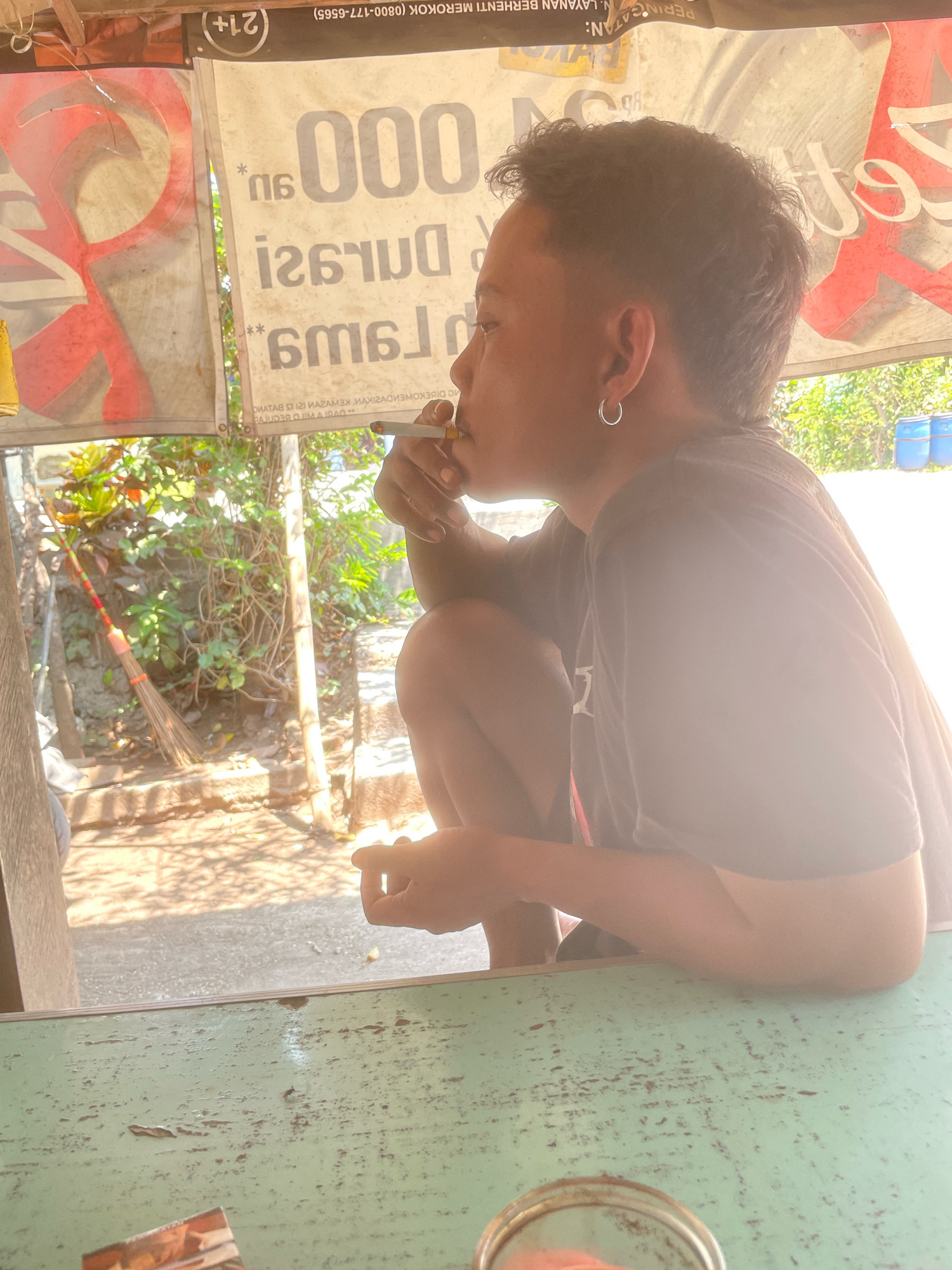 The image depicts a young person smoking a cigarette while sitting at a green, weathered table outdoors. They are wearing a dark t-shirt and a silver hoop earring. Behind them, a white banner with red lettering and a red graphic is visible. The background shows lush greenery, a broom leaning against a pole, and blue barrels. The overall setting suggests a casual outdoor environment, possibly a street-side stall or shop in Kebon Waris, Indonesia, given the Indonesian text on the banner. The lighting indicates it is daytime, with strong sunlight creating dappled shadows.