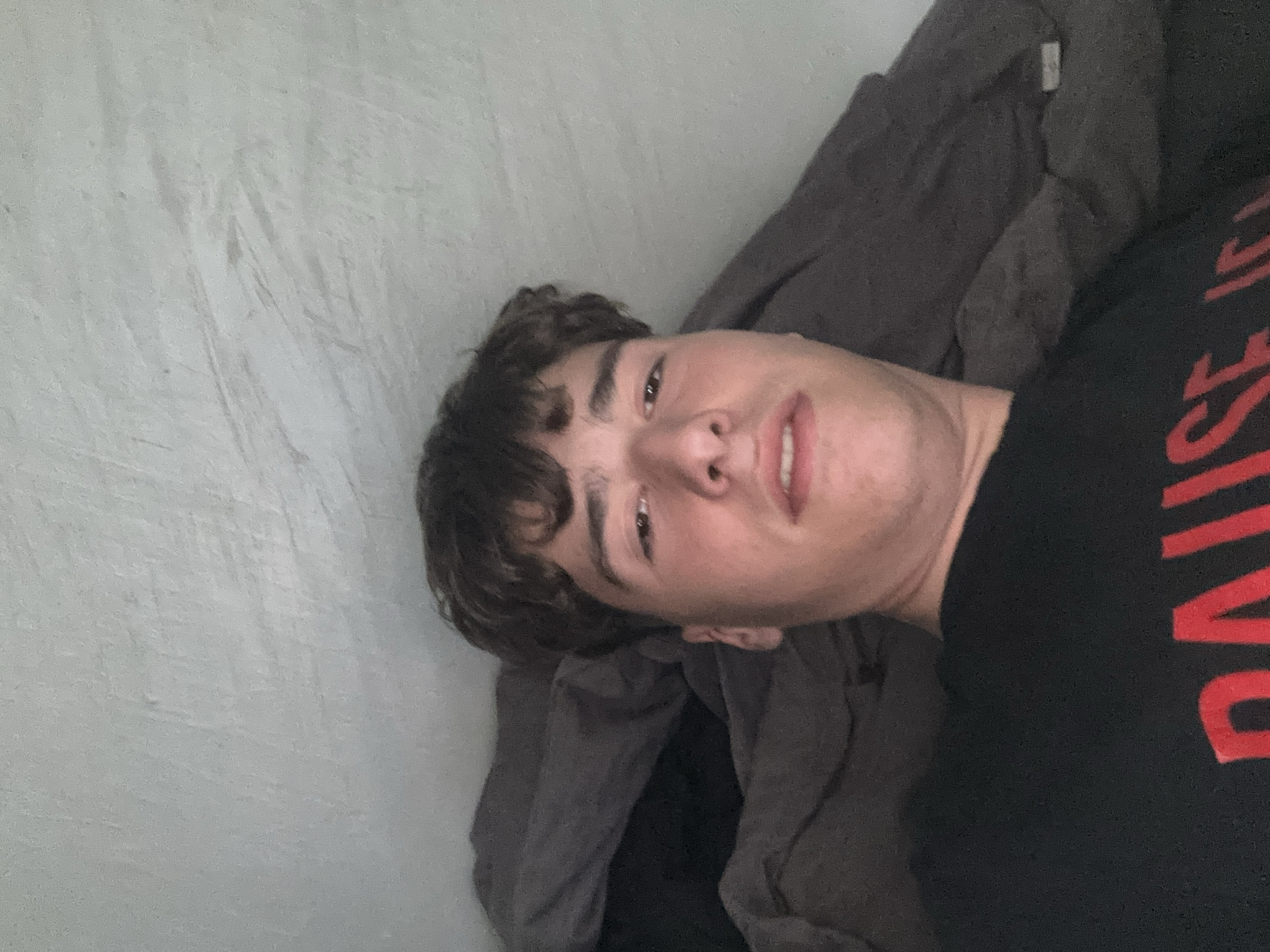 A young man with short, curly dark brown hair and brown eyes is featured in a close-up, rotated image, possibly a selfie. He is looking directly towards the camera with a slight, relaxed smile. He is wearing a black t-shirt with large, bright red text visible across the chest, which clearly spells "PAUSE". The background consists of a plain, light grey or off-white wall texture on the left, and a dark grey blanket or fabric visible behind and to his right. The man appears to be lying down or reclining against the fabric. The setting is indoors, under soft, ambient lighting. No specific location cues are visible within the image, though the context provided places it in Troyan, Bulgaria. The overall mood is casual and calm.
