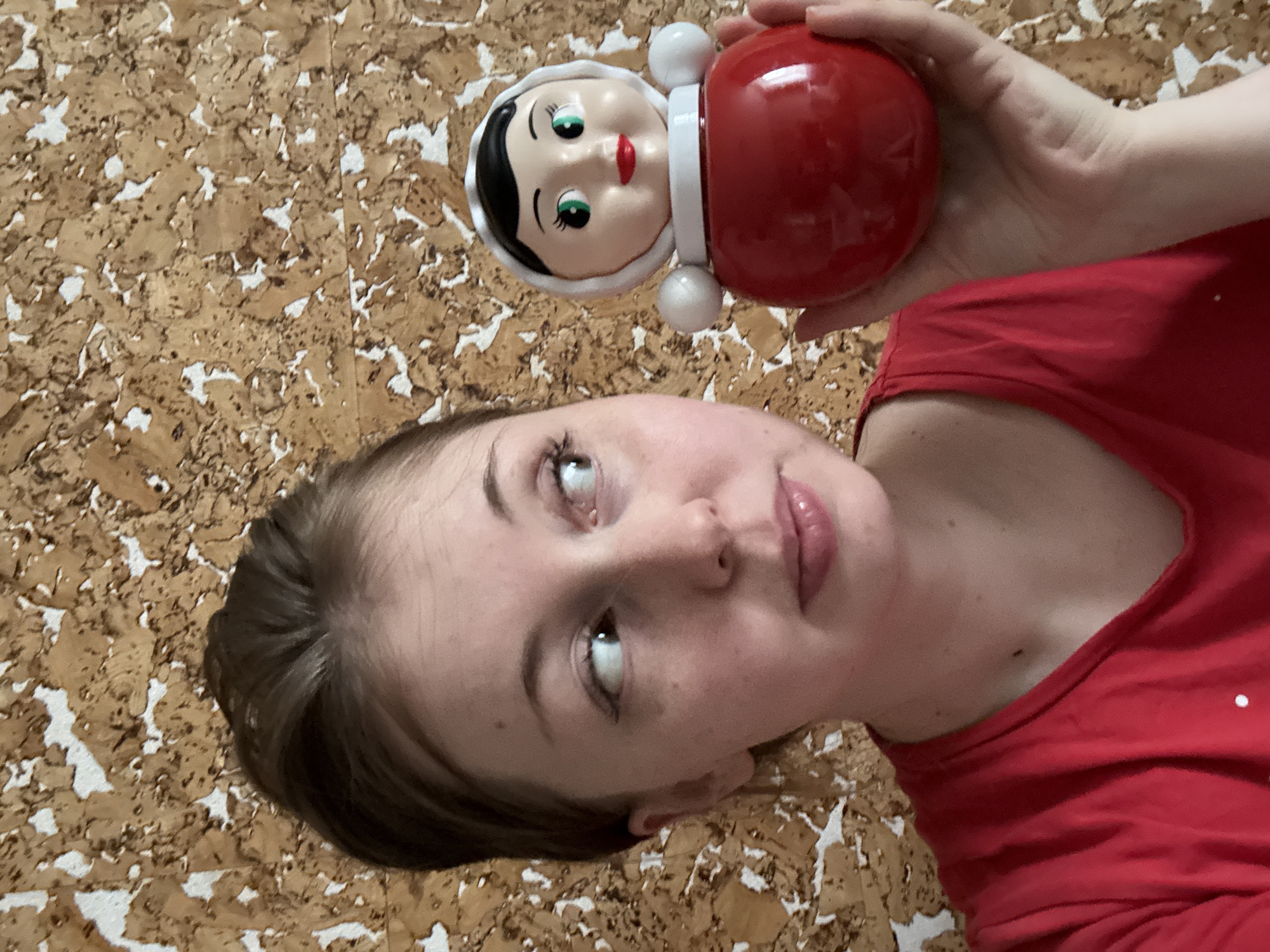 A woman is holding a roly-poly toy in front of a wall covered with cork board. The woman is wearing a red shirt and looking upwards and to the left. The roly-poly toy has a red body and a white face with black hair and green eyes. The background is a cork board with brown and white patterns. The location is Unknown, Unknown.