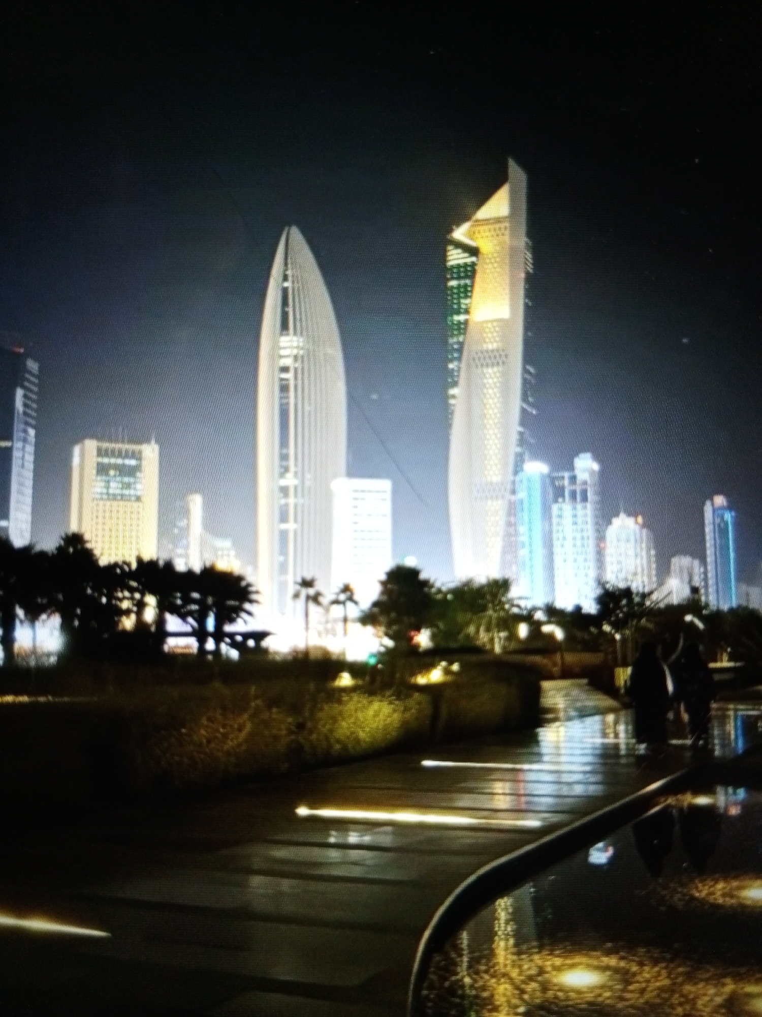The image depicts a nighttime view of a modern cityscape, with several tall skyscrapers illuminated against a dark sky. Two prominent, uniquely shaped skyscrapers dominate the center of the frame. The one on the left is a tall, slender, white building with a pointed, tapering top. The skyscraper on the right is also tall, with a distinctive angular design, featuring a golden-colored illuminated section near its peak and green lights further down its facade.
In the foreground, there is a park-like area with silhouetted trees, including palm trees, and lush vegetation. The ground appears to be wet, reflecting the city lights and creating streaks of illumination on the pathway. Several blurred figures of people can be seen walking in the middle ground, their forms indistinct due to the low light and motion. The overall atmosphere is one of urban development and modern architecture at night. The location is identified as Golbembal, Nigeria.