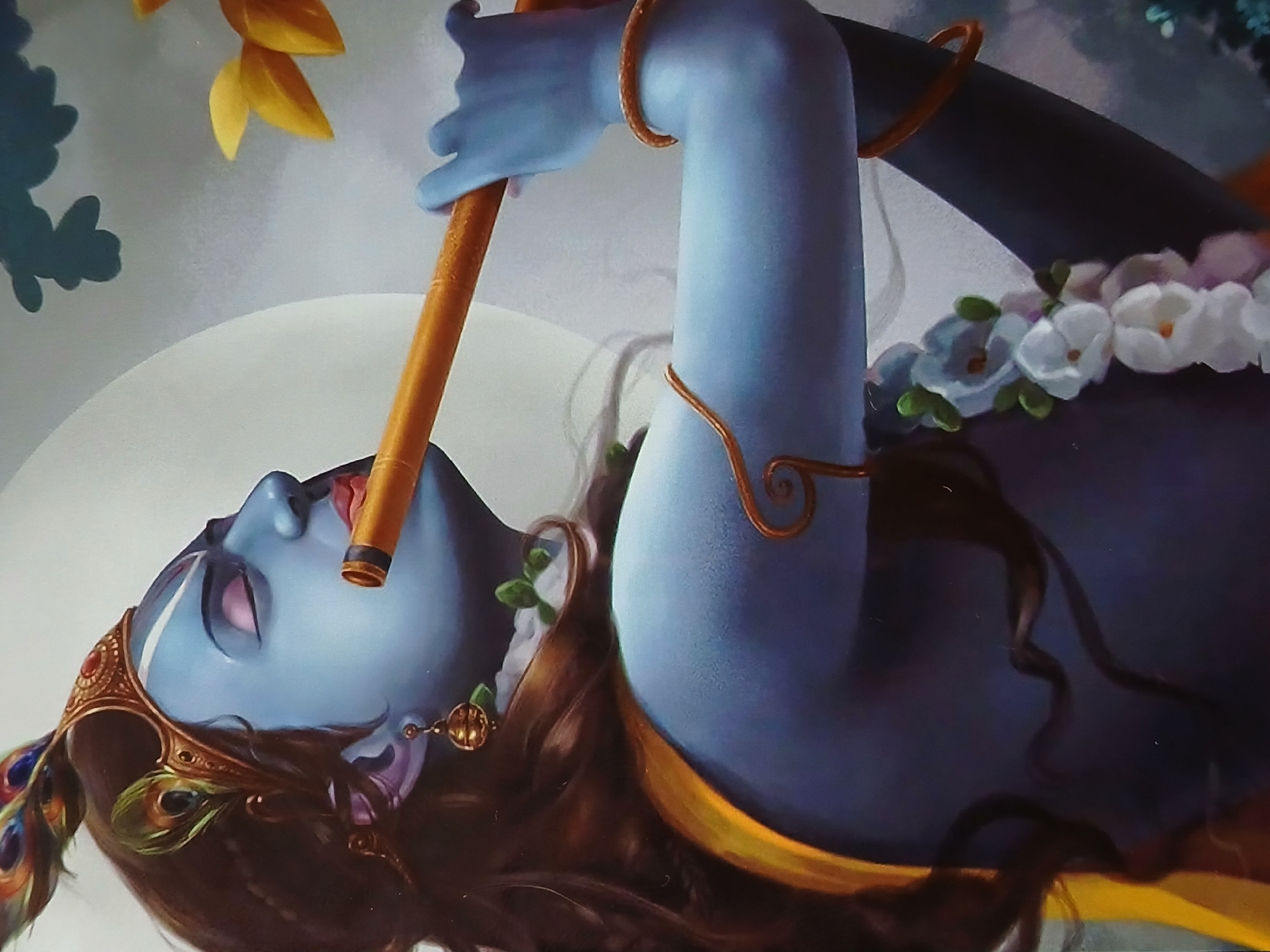 An illustration depicting the Hindu deity Krishna is presented. Krishna, with blue skin, is reclining with his head tilted back, eyes closed, and playing a flute. He is adorned with a golden crown featuring peacock feathers. His hair is long and dark brown. A golden armlet is visible on his upper arm, and he wears a garland of white flowers around his neck. The background features a white circular shape, possibly representing the moon, and green foliage in the upper left corner. The location of this image in Bengaluru, India cannot be determined from visual data alone.