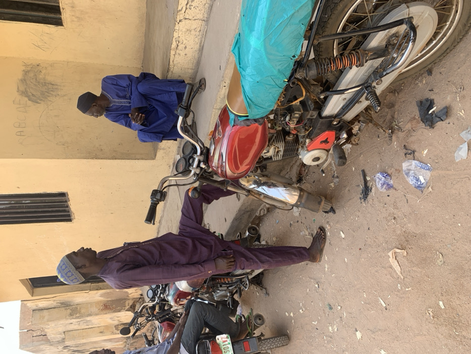The scene depicts a street in Bojude, Nigeria. Three adult males and multiple motorcycles are present on an unpaved, dusty ground with scattered litter, including plastic bags and dark debris. In the foreground, a red motorcycle with chrome accents and visible engine components is positioned. Its seat is covered with a turquoise fabric, and the front fender is missing. A male individual, wearing a purple long-sleeved tunic and trousers with a patterned cap and dark flip-flops, stands alongside this motorcycle, his right foot resting on its handlebar. He faces right in profile, looking slightly upwards. To the left, another male is partially visible, seated on a different motorcycle with a partially visible license plate ending "S01VZ". In the background, against a light yellow building with two windows, a third male sits on a raised concrete step. He wears a dark blue traditional garment with embroidery and a dark cap, with his hands clasped in front of him, looking towards the left. Faint "ABCDE" graffiti is visible on the yellow building's wall. An older, cracked building is partially visible to the far left. The scene is brightly lit by natural light.