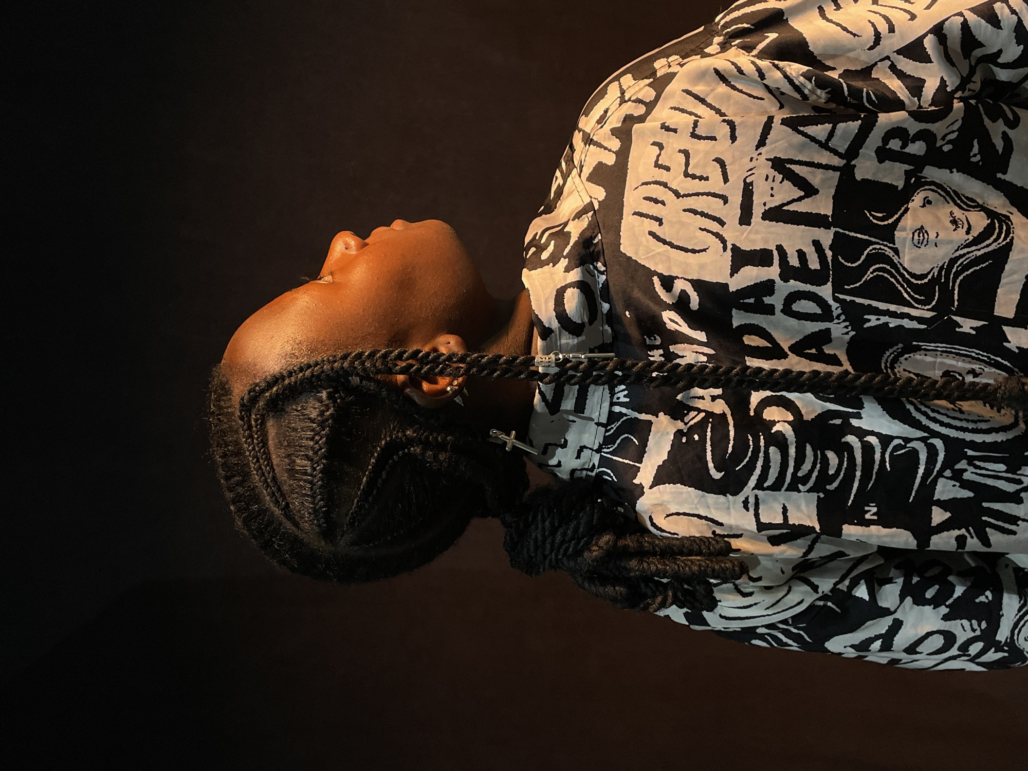 A medium shot depicts a Black person with dark brown skin, braids, and a silver cross earring, against a dark background. The person's head is tilted back, revealing a slight view of their face and neck. They are wearing a black and white patterned jacket. The lighting appears to be focused on the person. It is not possible to determine the location as Barakin Akawo, Nigeria from the image.