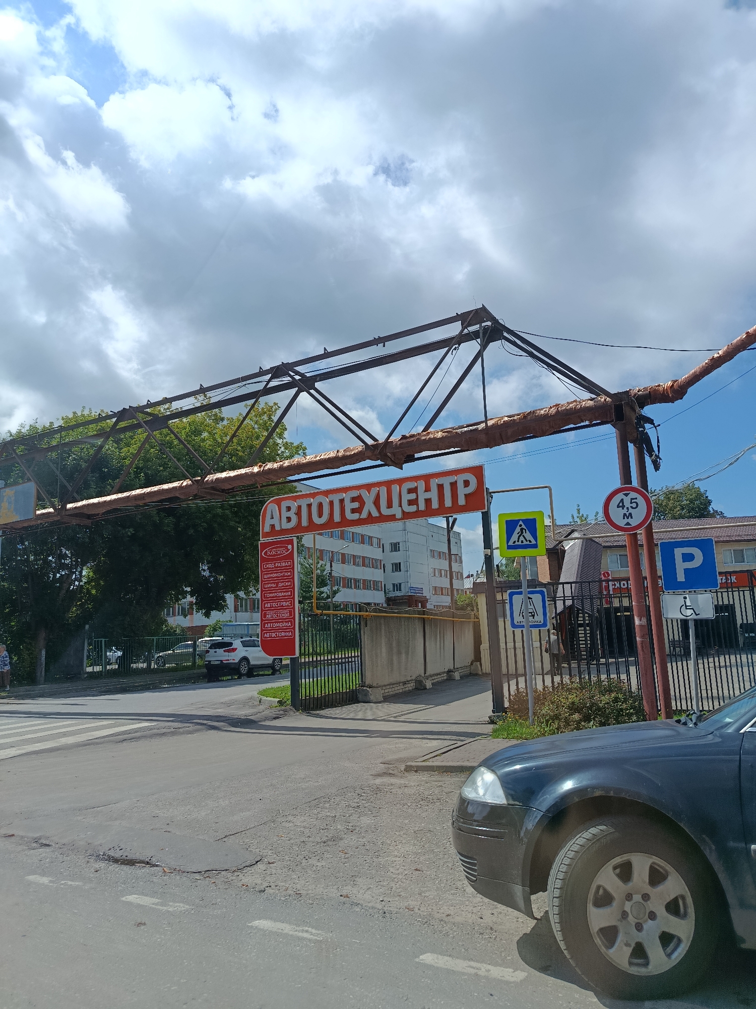 A street-level, slightly upward-angled photograph captures an overcast day in Orekhovo-Zuyevo, Russia. In the foreground, a black car is parked on a paved road. To the left, a crosswalk is visible. Further into the scene, an overhead metal structure, likely an overpass or support beam, spans the road. Attached to this structure is a large, orange sign with the Cyrillic text "АВТОТЕХЦЕНТР" (Autotechcenter). Beneath the main sign, a smaller, red sign lists various automotive services in white Cyrillic text, including "Космос," "Сход-развал," "Шиномонтаж," "Шины. Диски," "Тонировка," "Автосервис," "Автостекол," "Автомойка," and "Автостоянка."
To the right of the main sign, several road signs are present. One circular sign indicates a height restriction of 4.5 meters. Another sign depicts a pedestrian crossing. A blue parking sign with a white "P" is also visible, along with a sign for accessible parking. In the background, a multi-story building with rows of windows stands behind a metal fence. Trees and greenery are abundant, suggesting a suburban or mixed urban/green area. The sky is filled with dramatic, grey clouds, hinting at potential rain. A person, possibly a child, is partially visible on the far left side of the image, standing near a fence. The overall impression is that of a functional, service-oriented area within a Russian city.