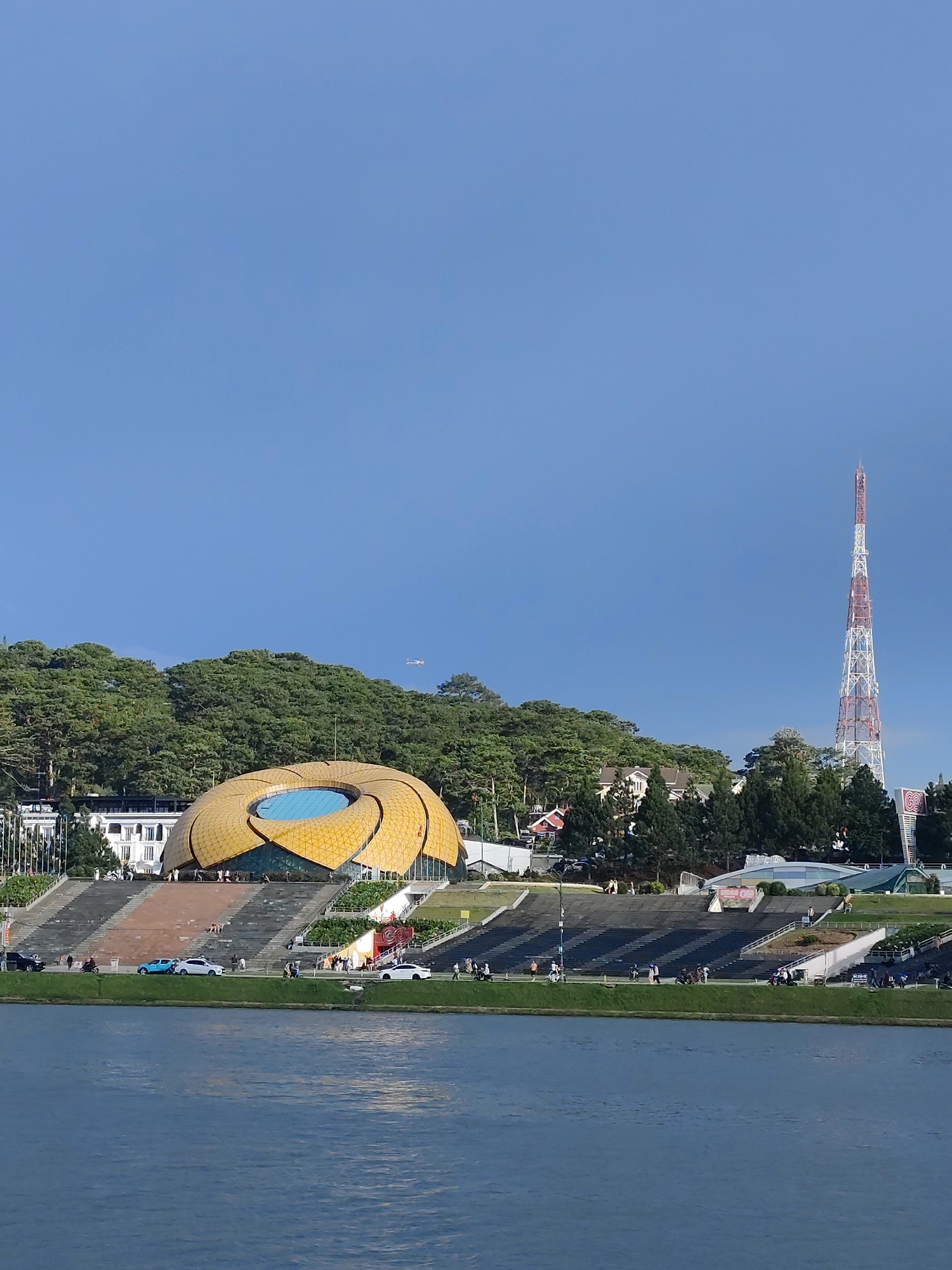 A wide-angle, eye-level, outdoor, vertical photograph captures a clear, bright blue sky above a bustling city scene. In the foreground, a calm, reflective blue body of water fills the bottom third of the frame. Beyond the water, a landscaped embankment is lined with a walking path and a set of wide, dark stairs leading up to a distinctive, golden, flower-shaped building with a central blue circular opening. The building is situated on a hillside covered with lush green trees. To the right of the flower building, a tall, red and white lattice radio tower pierces the sky. Several people are visible walking and gathering on the embankment and stairs. A few cars, including a blue sedan and a white sedan, are parked or driving along the path. A small, light-colored aircraft is visible as a tiny speck in the sky above the trees. The overall impression is one of a well-maintained public space with a mix of natural and architectural elements.