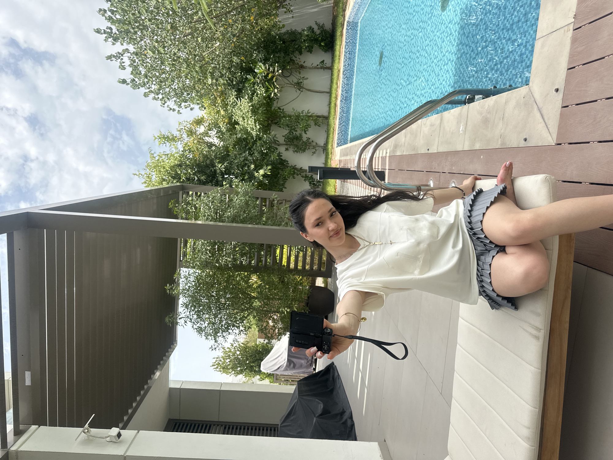 A woman is depicted lying or reclining on a white cushioned lounge chair outdoors, situated on a wooden deck adjacent to a swimming pool. She has long, dark hair and is wearing a light-colored short-sleeved top and a grey pleated skirt. In her right hand, she holds a compact digital camera, pointing it towards herself, appearing to take a selfie or record a video. Her feet are bare and resting on the end of the lounge chair.
The setting features a bright blue swimming pool with a metal ladder leading into it. Surrounding the pool and deck area is abundant green foliage, including trees and shrubs, suggesting a lush outdoor environment. In the background, modern architectural elements are visible, including a dark slatted structure or wall and a white overhead covering. The sky above is bright with scattered white clouds, indicating daytime. A security camera is mounted on a white wall in the background. The scene suggests a relaxed moment in a sunny, upscale outdoor location, consistent with the provided context of Dubai, United Arab Emirates. No discernible text is visible.