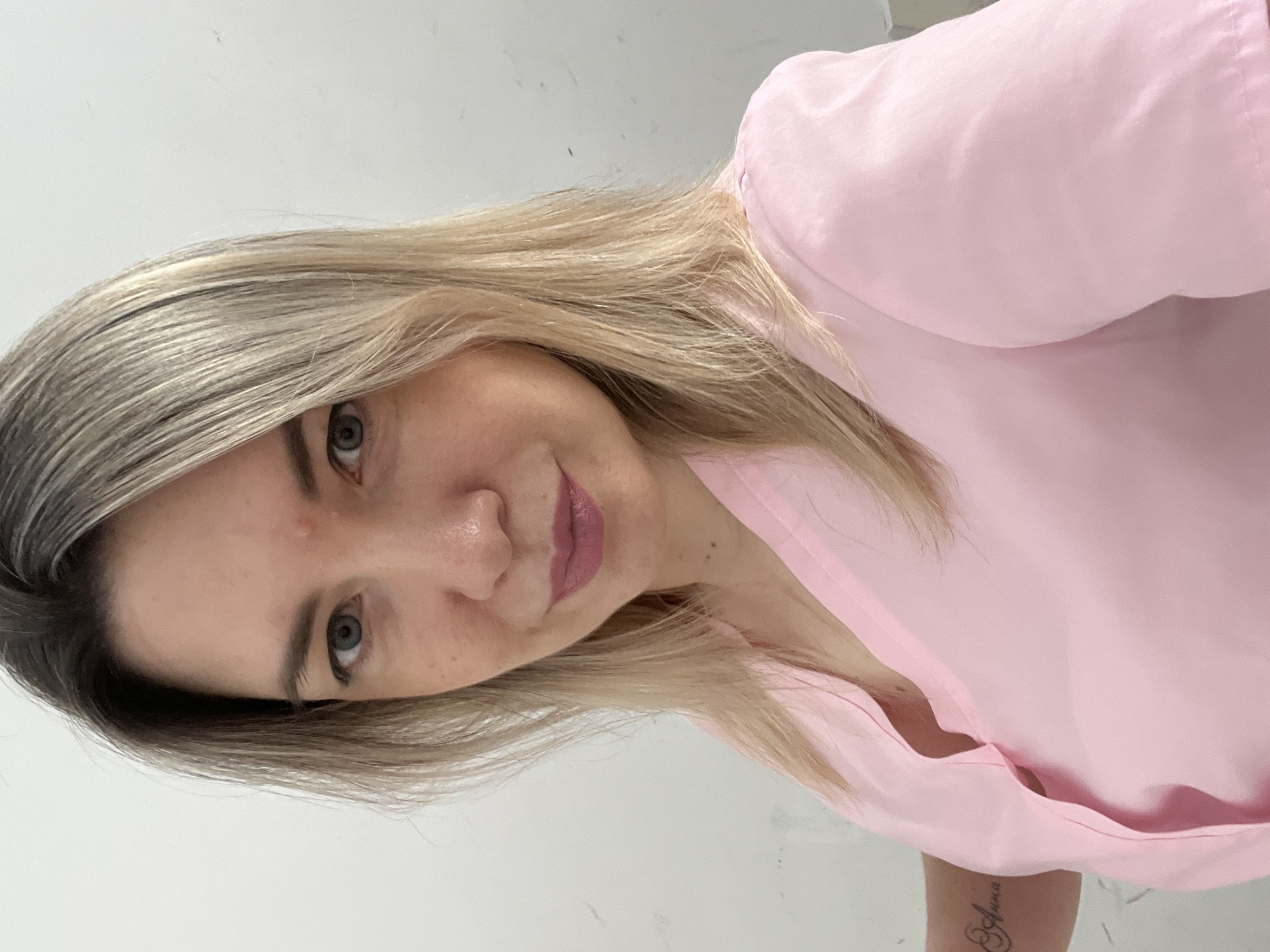 A woman with blonde hair and green eyes, wearing a pink v-neck shirt, is pictured from the chest up against a white wall. She has a tattoo on her left arm that reads "Anna." The background is a plain white wall, with no discernible city or environmental details to suggest a specific location in Sochi, Russia, or elsewhere.