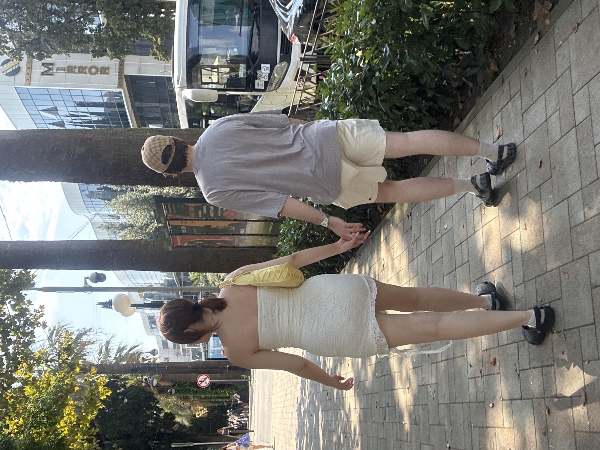 A couple, a woman and a man, walk hand-in-hand down a sun-dappled sidewalk in Sochi, Russia. The woman, facing away from the camera, wears a white strapless lace dress and carries a yellow purse over her shoulder. She has her hair pulled back in a ponytail. The man, also facing away, wears a light grey t-shirt, beige shorts, a checkered baseball cap, and dark patterned clogs with white socks. They are walking on a paved sidewalk lined with trees and bushes. In the background, a modern building with reflective glass windows and the word "MIRROR" visible is situated across the street, alongside a white bus and a dark car. The setting appears to be a public area with signage and streetlights.