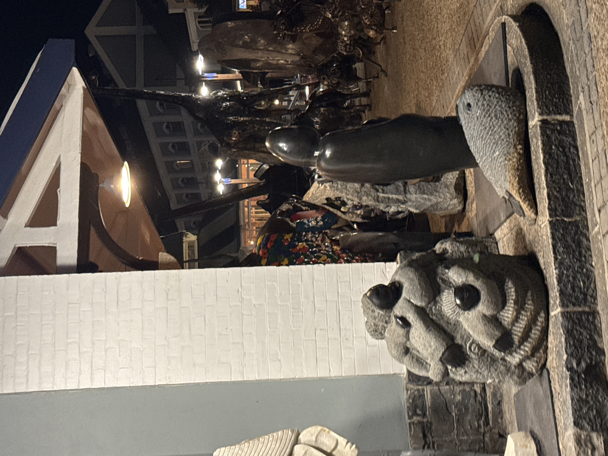 The image depicts a dimly lit street scene in Cape Town, South Africa, at night. A white building with a blue-trimmed, triangular gable is visible. In the foreground are various stone sculptures: a stylized fish head emerging from an arched frame, smooth elongated shapes resembling seals, and a complex carving of a stylized face with dark, prominent eyes. Behind the sculptures are other metal and wooden sculptures, with hints of people and colorful textiles in the background. A warm light shines from within the gable structure. The street appears to be paved with stone or brick.