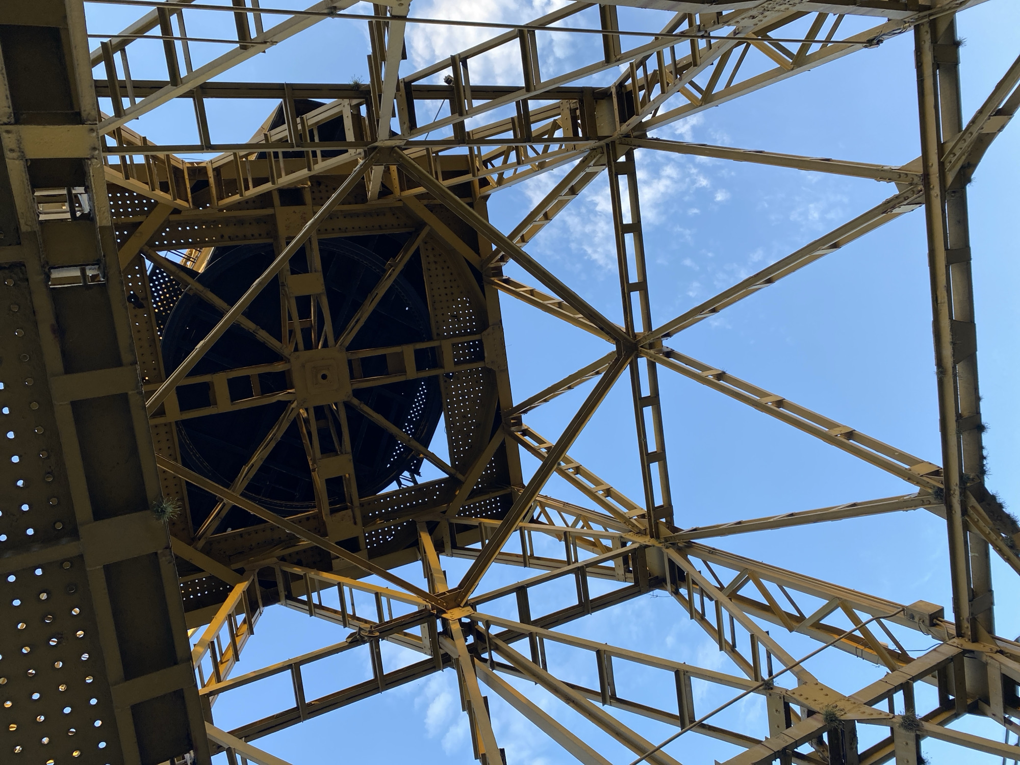 The image captures an upward view of a yellow metal structure against a blue sky with wispy clouds. The structure is comprised of interconnected beams and crossbars, forming a complex network. A large, dark, circular object, likely part of a mechanical apparatus, is integrated within the structure. There is some green vegetation growing from the metal. The location is likely in Buenos Aires, Argentina, given the style of architecture.
