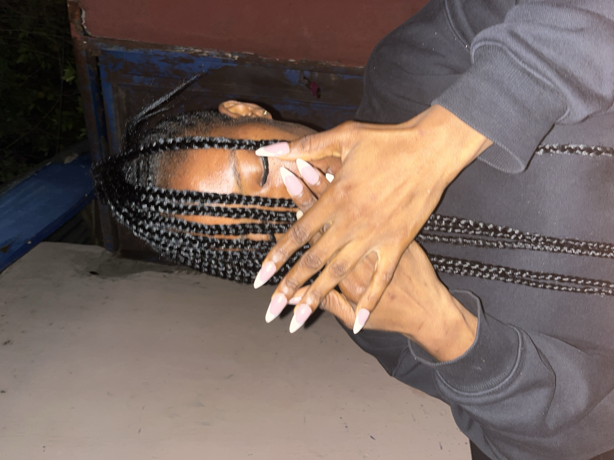 The image shows a close-up of a person's upper body, specifically their face partially obscured by their hands. The person has dark skin and braided hair. Their hands are positioned in front of their face, and they have long, light-colored fingernails with white tips. The person is wearing a dark-colored hooded sweatshirt. The background appears to be outdoors, with a textured wall on the left and a rusted metal structure on the right, suggesting an urban or semi-urban environment. The lighting is dim, indicating it might be evening or night. This scene could plausibly be located in Jos, Nigeria, given the commonality of such environments and the appearance of the individual.