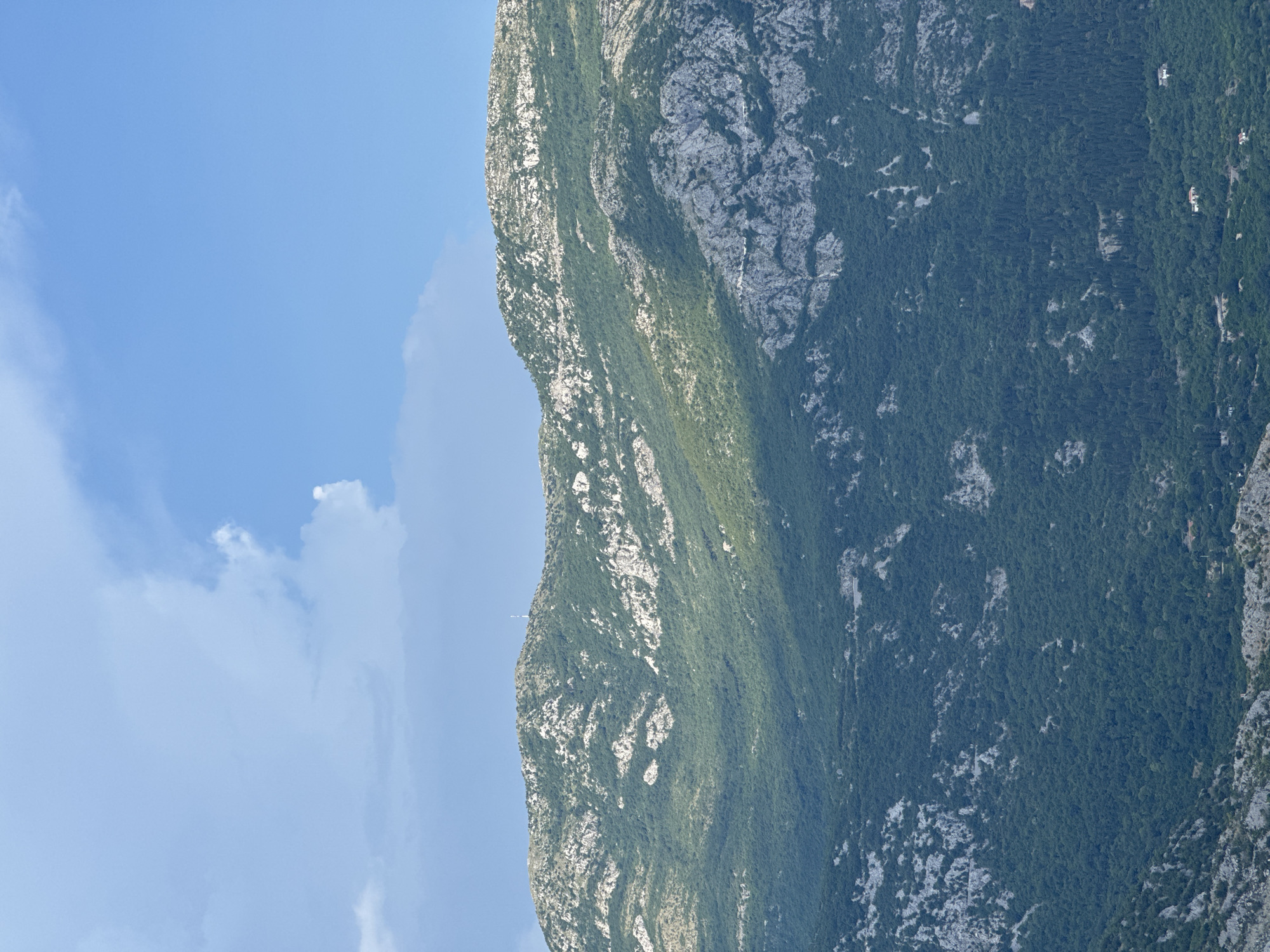 The image shows a view of a verdant mountainside in Štrp, Montenegro, under a bright blue sky with scattered clouds. The mountain is covered in dense green foliage, interspersed with patches of rocky, lighter-colored terrain. Sunlight casts shadows across the slopes, highlighting the undulating contours of the landscape. A thin antenna or tower is visible near the summit of the mountain. The lower portion of the image reveals a scattering of houses nestled among the trees at the base of the mountain.