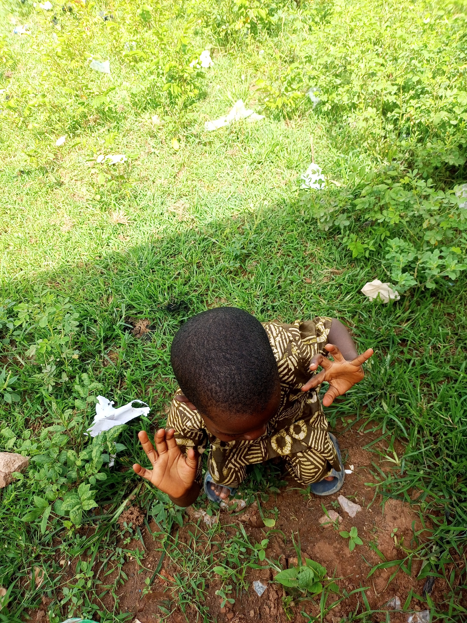 A child, facing away from the camera, is crouched on the ground in a grassy area. The child is wearing a patterned brown and gold outfit and dark sandals. The child's hands are raised and open, palms facing upwards, with fingers spread. The ground is covered in green grass, some dirt, and scattered pieces of white paper. The scene appears to be outdoors, possibly in a residential area or park. The overall lighting suggests it is daytime, with shadows cast by the sun. The location is specified as Tempure, Nigeria.