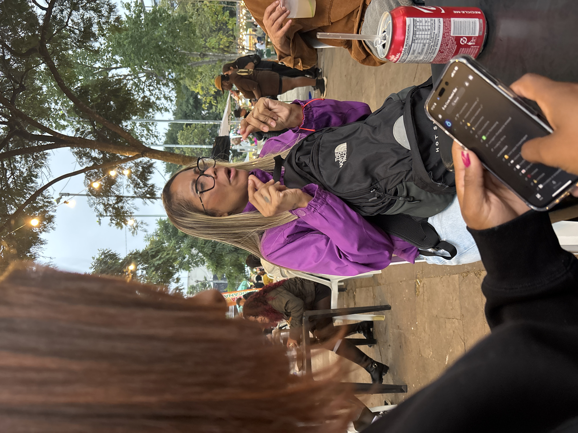 Here is a description of the image.
The image depicts a group of people gathered at an outdoor dining area, likely a street-side cafe or restaurant. The location suggests a lively urban atmosphere.
A woman with glasses and blonde hair, wearing a purple jacket and a black backpack, is seated at a table. She seems to be talking and gesturing with her hands. A person in a black sweatshirt holds a cell phone with pink painted nails. There is also a person with auburn hair sitting at a table.
Several objects are visible on the table, including a can of soda with a straw. Other people are sitting and walking in the background.
Based on the signs, the location appears to be in the city of São Paulo, Brazil. The outdoor setting is enhanced by string lights hanging above, creating a warm and inviting atmosphere.