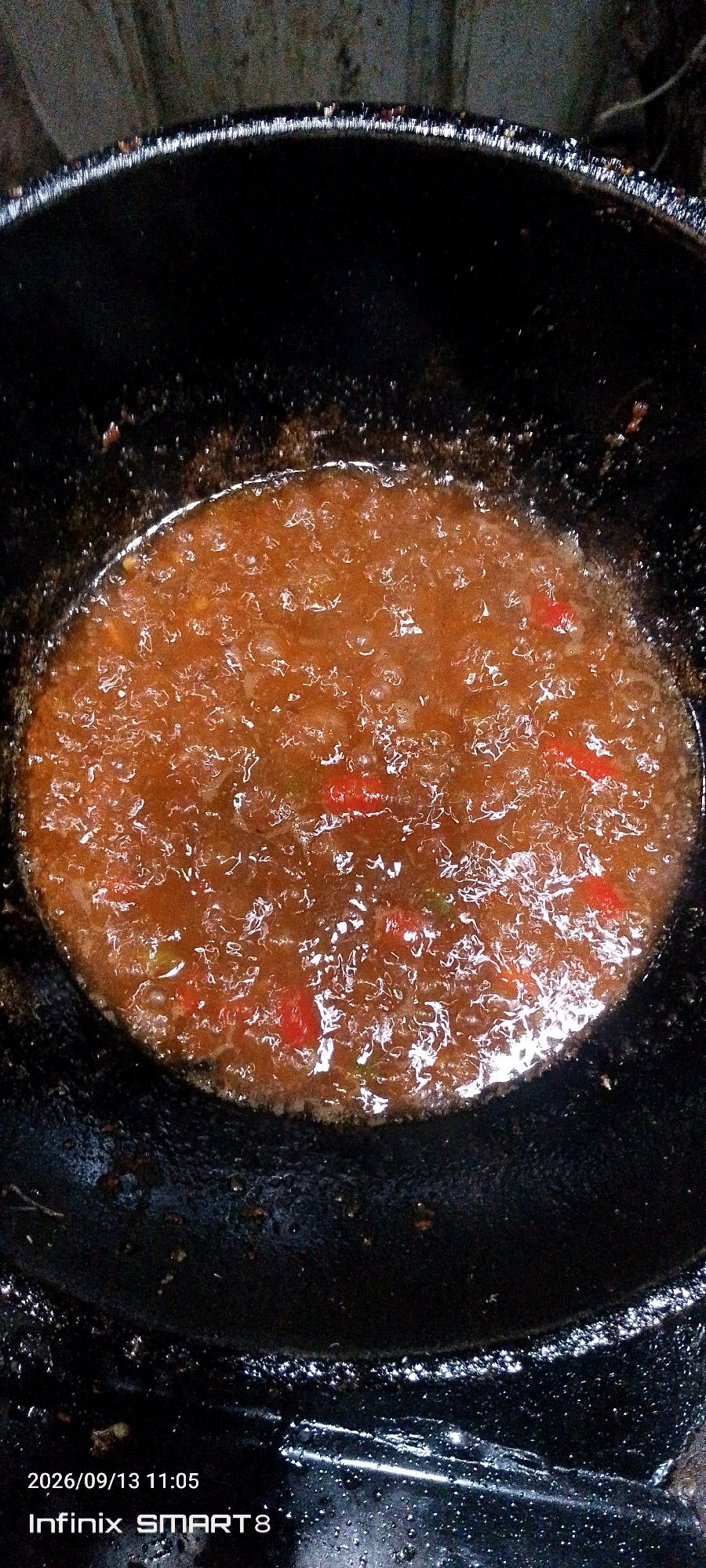 The image shows a close-up, overhead view of a dark, possibly cast-iron, cooking pot containing a bubbling, reddish-brown sauce. The sauce appears to have small pieces of food, possibly vegetables or meat, and some red flecks, which could be chili peppers. The pot's rim is visible, and the background is dark and out of focus, suggesting a dimly lit environment. The bottom right corner of the image displays a timestamp "2026/09/13 11:05" and text "Infinix SMART 8". There is no indication of the city of Serang, Indonesia, within the visual content of the image.
