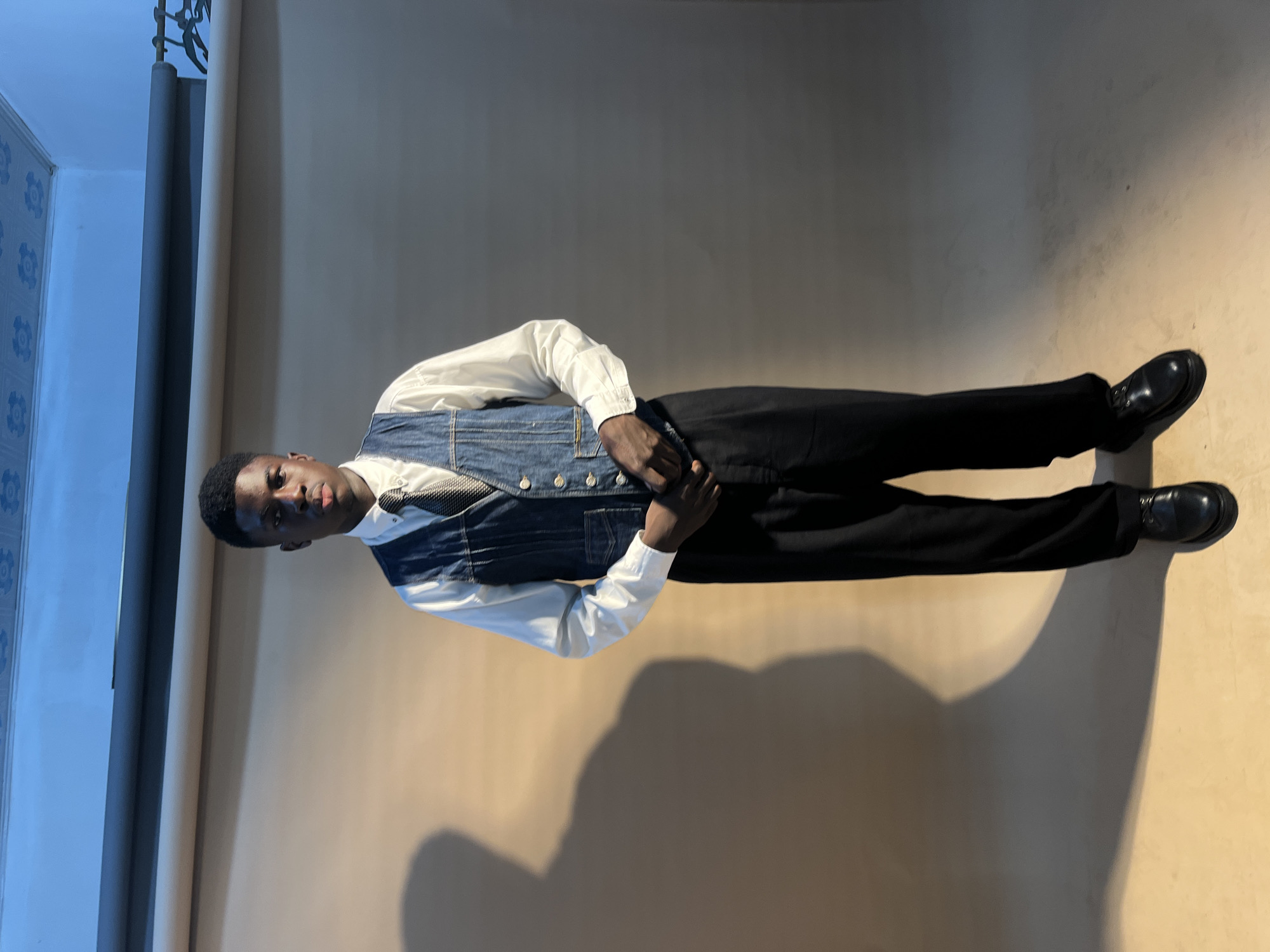 A young Black man is standing in a brightly lit indoor location. He is wearing a white long-sleeve shirt, a denim vest, black pants, and black leather shoes. He is looking towards the camera with his hands clasped together. The background is a combination of blue wall/corner and tan walls. The location is Gura Top, Nigeria.