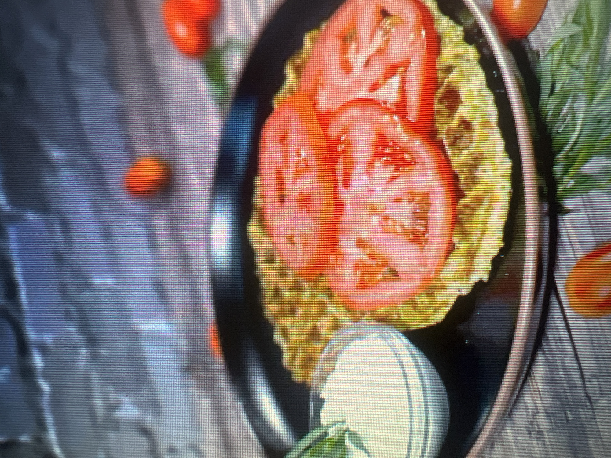 A close-up shot shows a savory waffle topped with three slices of red tomato and served on a black plate. To the left of the waffle is a small glass bowl containing a white dip, possibly sour cream or yogurt, with a few sprigs of green basil. The waffle appears to be golden brown and has a grid pattern. In the background, there are scattered cherry tomatoes and more basil leaves. The entire scene is set against a textured, dark surface that resembles a wooden table. The image appears to be a photograph of food, likely taken in a studio or kitchen setting. The location is Unknown, Unknown.