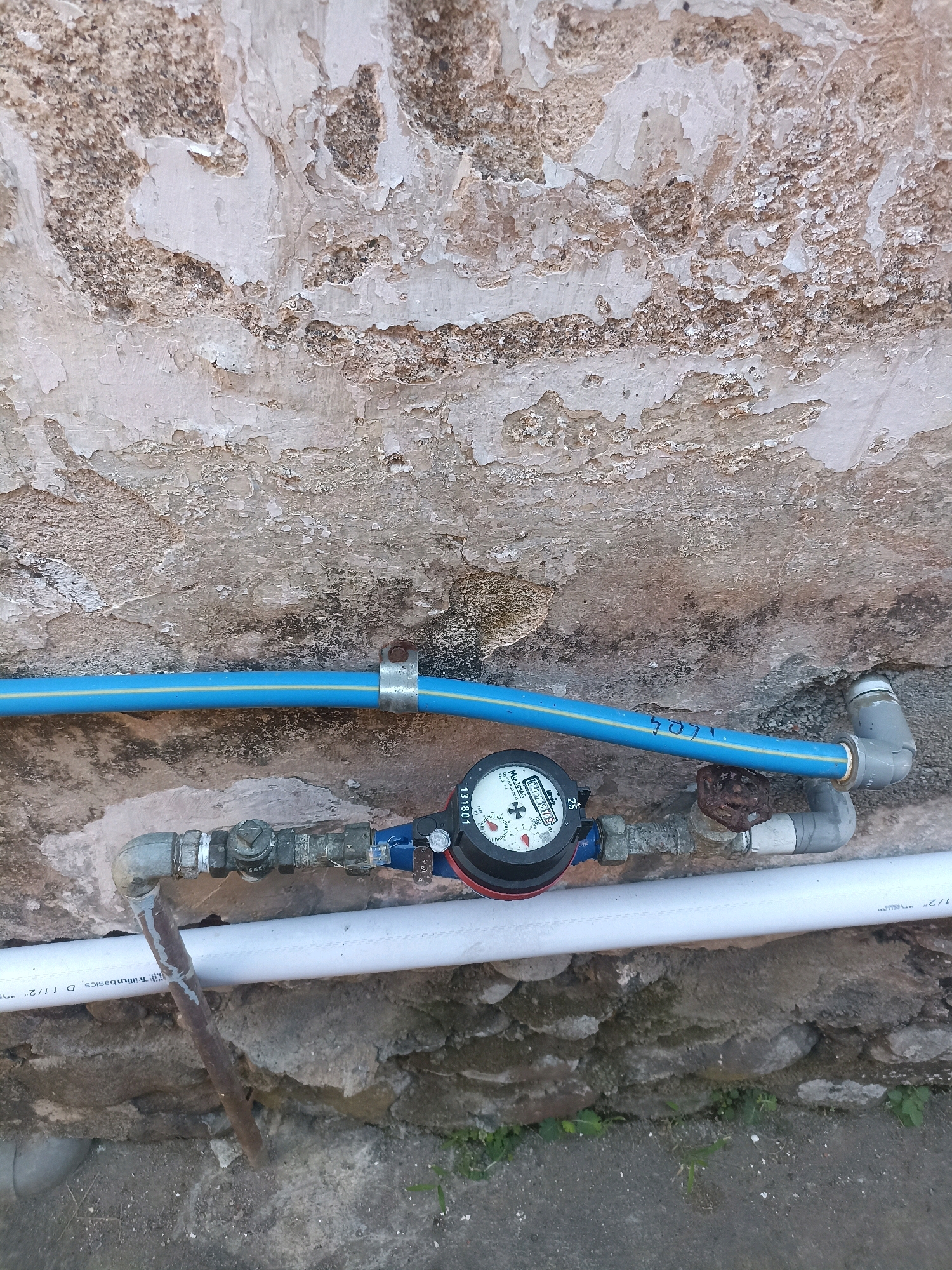 The image shows a close-up of a water meter installed on pipes attached to a weathered, textured wall. The wall, which appears to be made of concrete or stucco with peeling paint, occupies the upper portion of the frame. Below the wall, a blue pipe runs horizontally, secured by a metal clamp. Attached to this pipe is a water meter with a circular face displaying readings. Below the water meter, a network of pipes and fittings is visible, including a shut-off valve and connections to other pipes. A white pipe runs horizontally below the water meter and its associated fittings. A rusty metal rod protrudes from the ground near the pipes. The ground beneath the pipes appears to be rough and rocky, with some sparse green vegetation. The scene is outdoors, likely in an unmaintained area. The presence of the water meter suggests a residential or utility area, and the overall aesthetic points to a location where infrastructure is exposed to the elements. The scene is static, with no people or animals present. The lighting suggests it is daytime, with no harsh shadows.