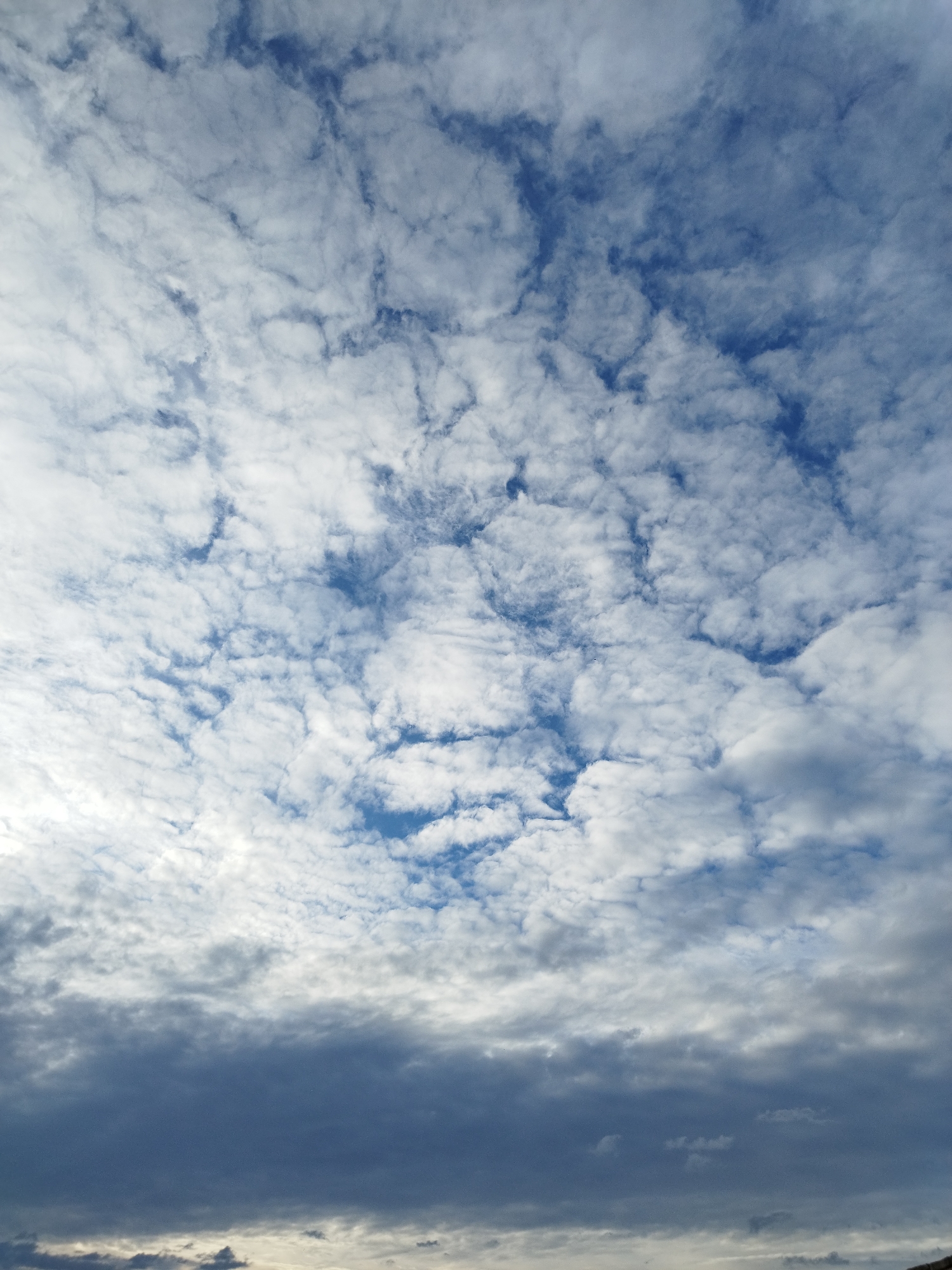 The image shows a sky filled with scattered clouds over Băleni-Sârbi, Romania. The clouds are predominantly white and grey, with patches of deep blue sky visible between them. The lower portion of the image features darker, more ominous-looking clouds, suggesting a change in weather. The overall impression is of a dynamic and natural sky. No people, subjects, objects, or actions are discernible in the image.