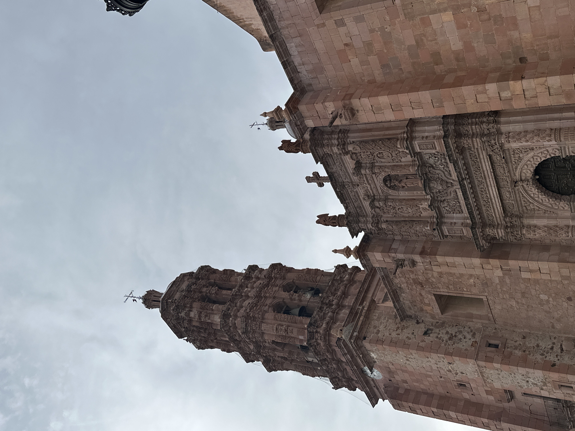 The image shows a low-angle view of the exterior of the Zacatecas Cathedral in Zacatecas City, Mexico, under an overcast sky. The cathedral is constructed of pinkish-red stone. A tall, ornate bell tower, also made of stone, dominates the left side of the frame. The tower features multiple levels with arched openings where bells are visible, intricate carvings, and a cross at its apex. A clock face is also visible on the lower part of the tower. To the right of the tower is the main facade of the cathedral, characterized by elaborate baroque carvings, niches with statues, and arched doorways. The building's facade is also made of the same reddish stone. The overall impression is of a grand and historically significant religious structure.