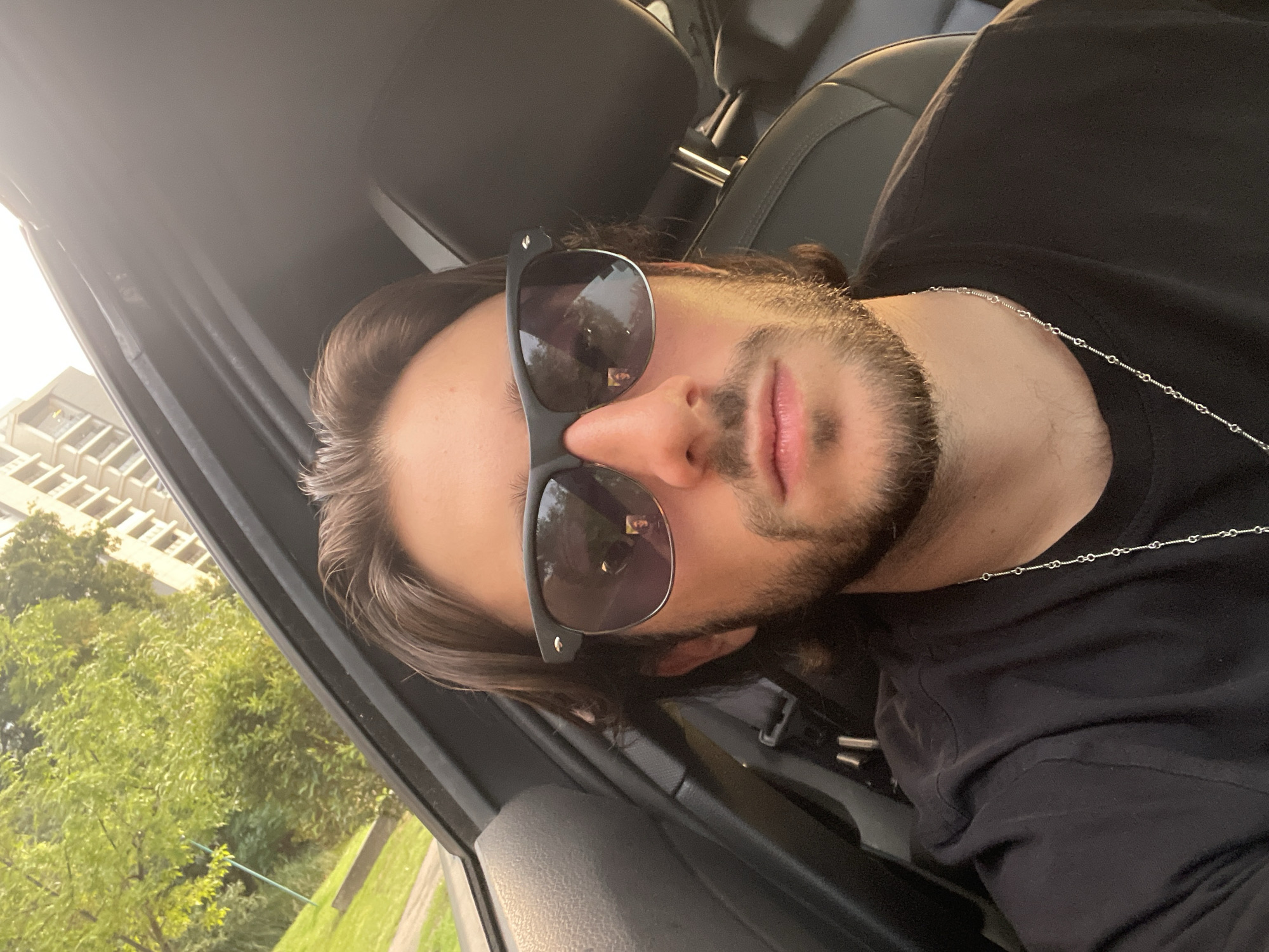 A selfie shot from inside a car shows a person with medium-length brown hair, a beard, and sunglasses. The person is wearing a black T-shirt and a silver chain necklace. The background visible through the car window includes green trees and a tall building. Reflections in the sunglasses show additional details, potentially indicating the location. Based on the general appearance and surrounding environment, the location is likely within Huixquilucan de Degollado, Mexico.